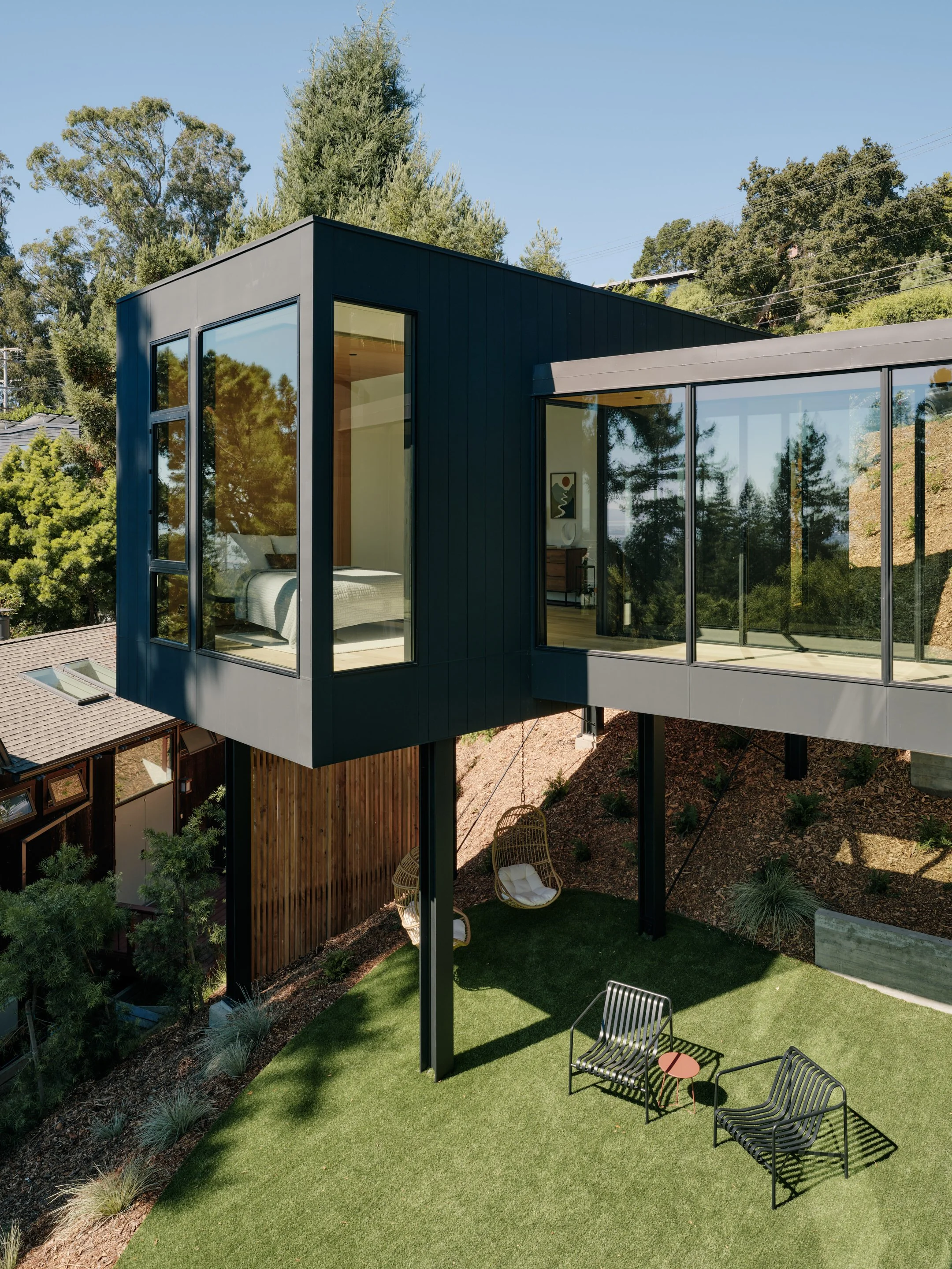 Modern house with large glass windows situated on a hillside, surrounded by greenery and trees, with a balcony and outdoor seating area beneath.