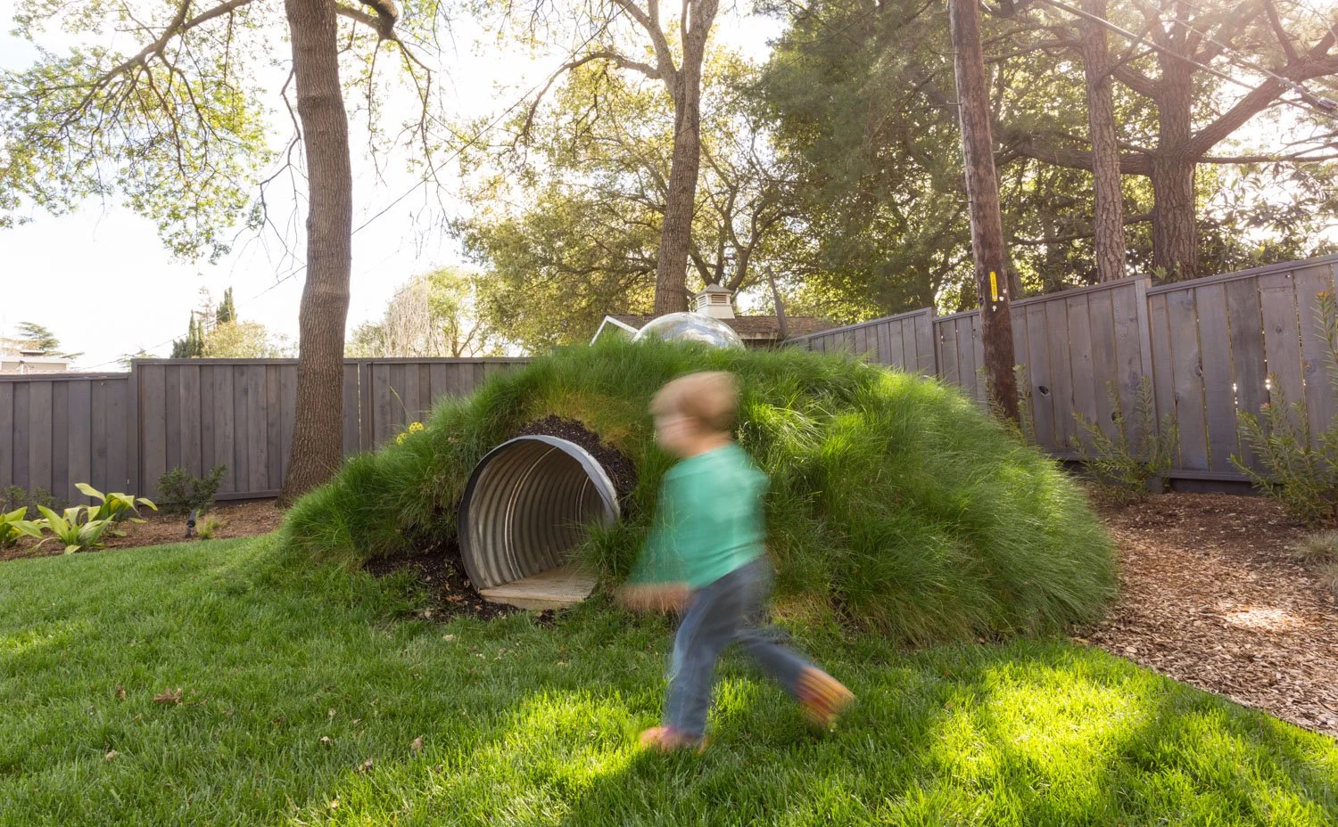 A child running in a backyard near a grass-covered tunnel-like play structure, with trees and a wooden fence in the background.
