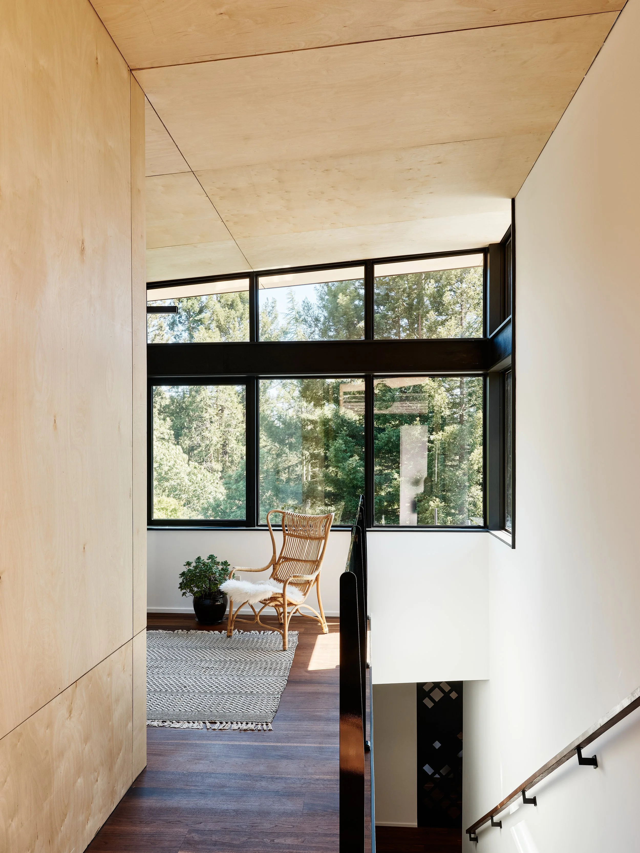 Interior of a modern home with large windows revealing trees outside, featuring a wicker chair with a white fur cushion, a small potted plant on the floor, and a woven rug on dark wooden flooring.