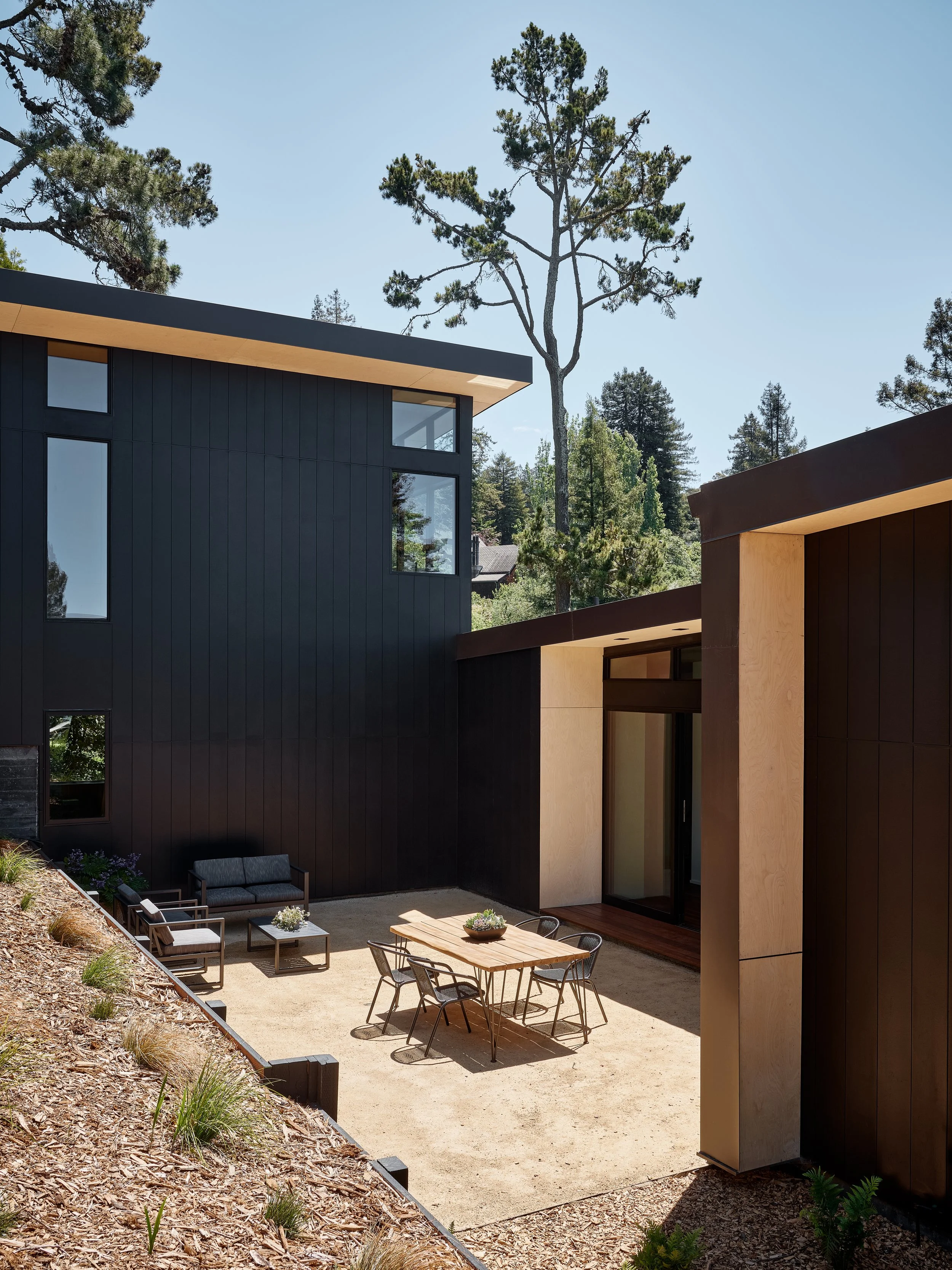Modern black and wood building with outdoor patio featuring seating, table, and surrounding trees.