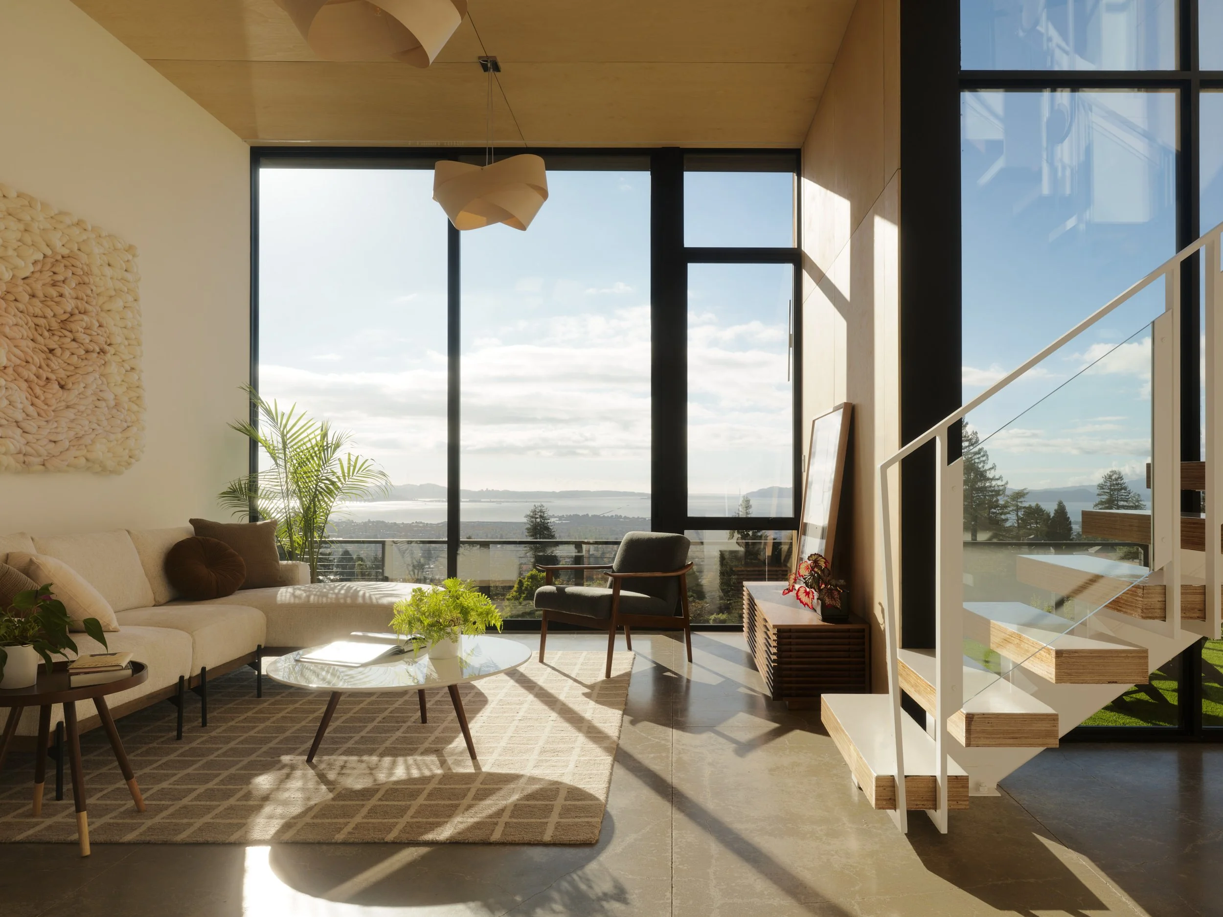 Modern living room with large floor-to-ceiling windows showing a view of water and trees outside, with a beige sofa, wooden chairs, a coffee table, and decorative plants inside.