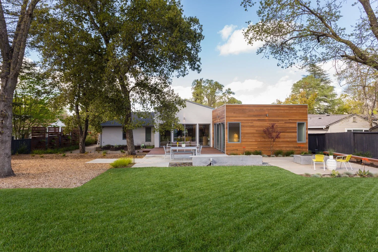 Modern house with a combination of white and wood exterior, surrounded by trees and a well-maintained lawn with outdoor furniture in the backyard.