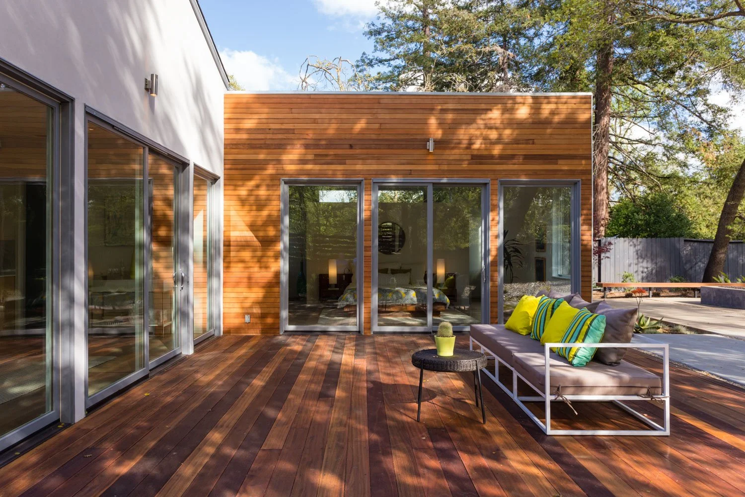 Modern house with large glass sliding doors leading to a wooden deck outdoor seating area with a cushioned bench and colorful pillows. The house features a combination of white and wood panel exterior, and the background shows tall trees and a sunny 