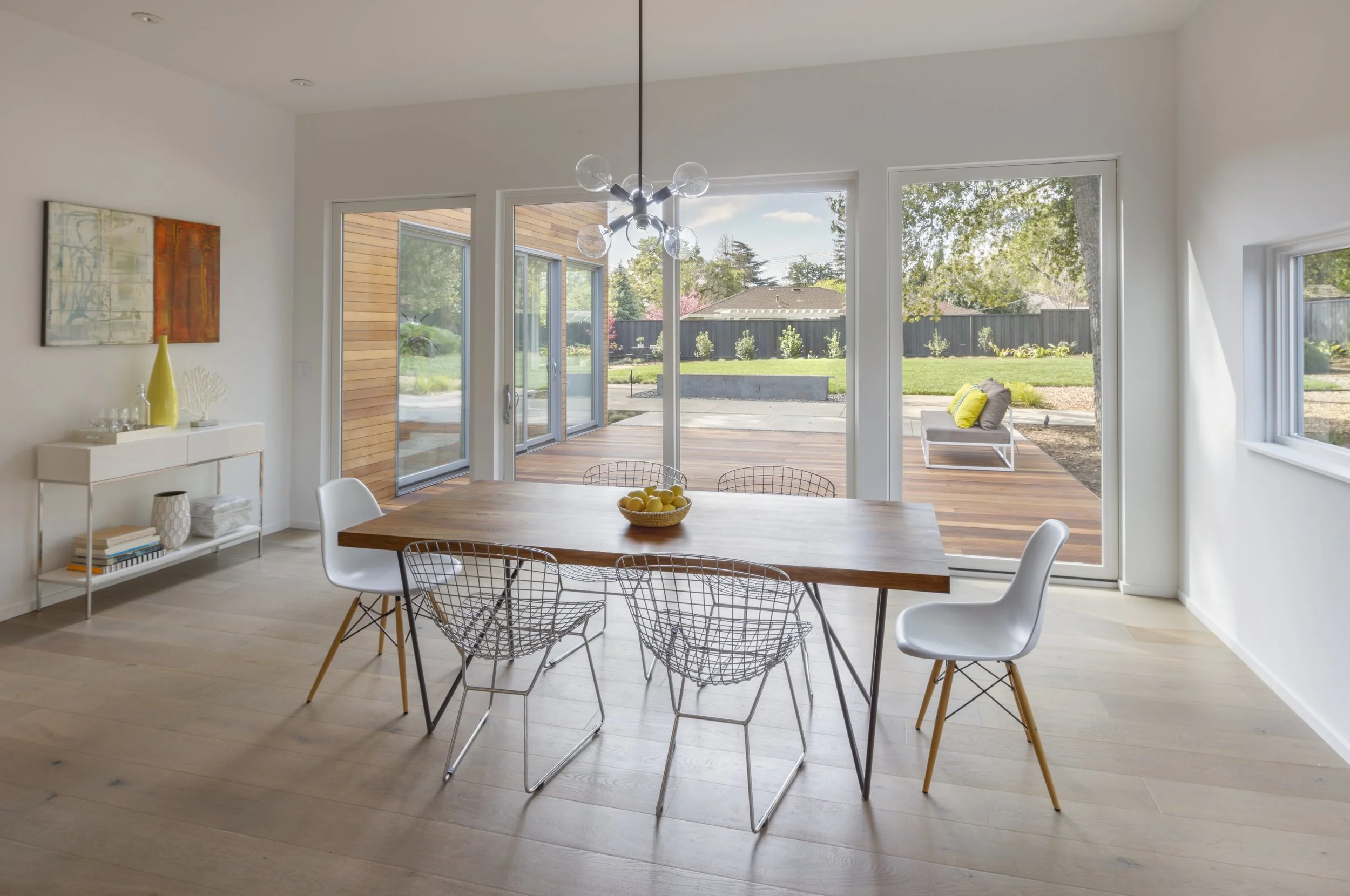 Interior view of a dining room with a wooden table, white chairs, a modern chandelier, and large sliding glass doors leading to a backyard with a patio and lawn.