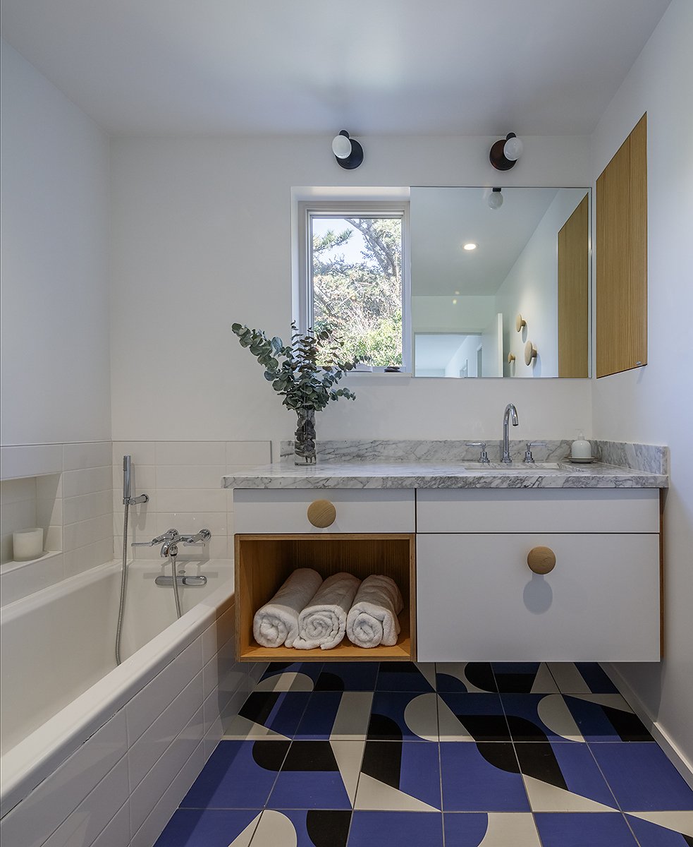 A modern bathroom featuring a bathtub, a sink with a marble countertop, a mirror, a window, and a cabinet with rolled towels inside. The floor has geometric black, white, and blue tiles.