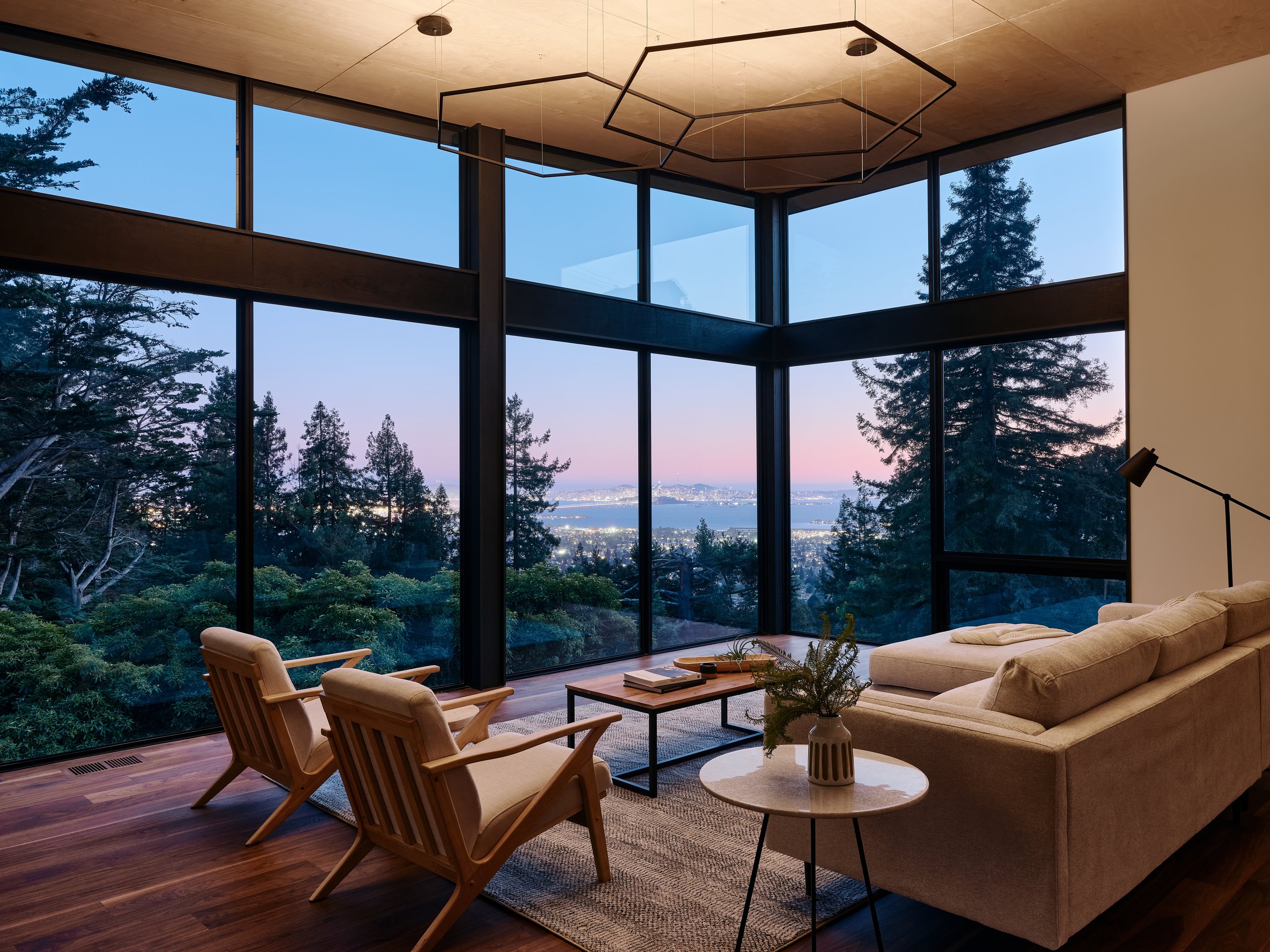 Modern living room with large floor-to-ceiling windows showing lush greenery outside. Features include a beige sofa, wooden chairs, a coffee table, decorative vases, and abstract wall art.