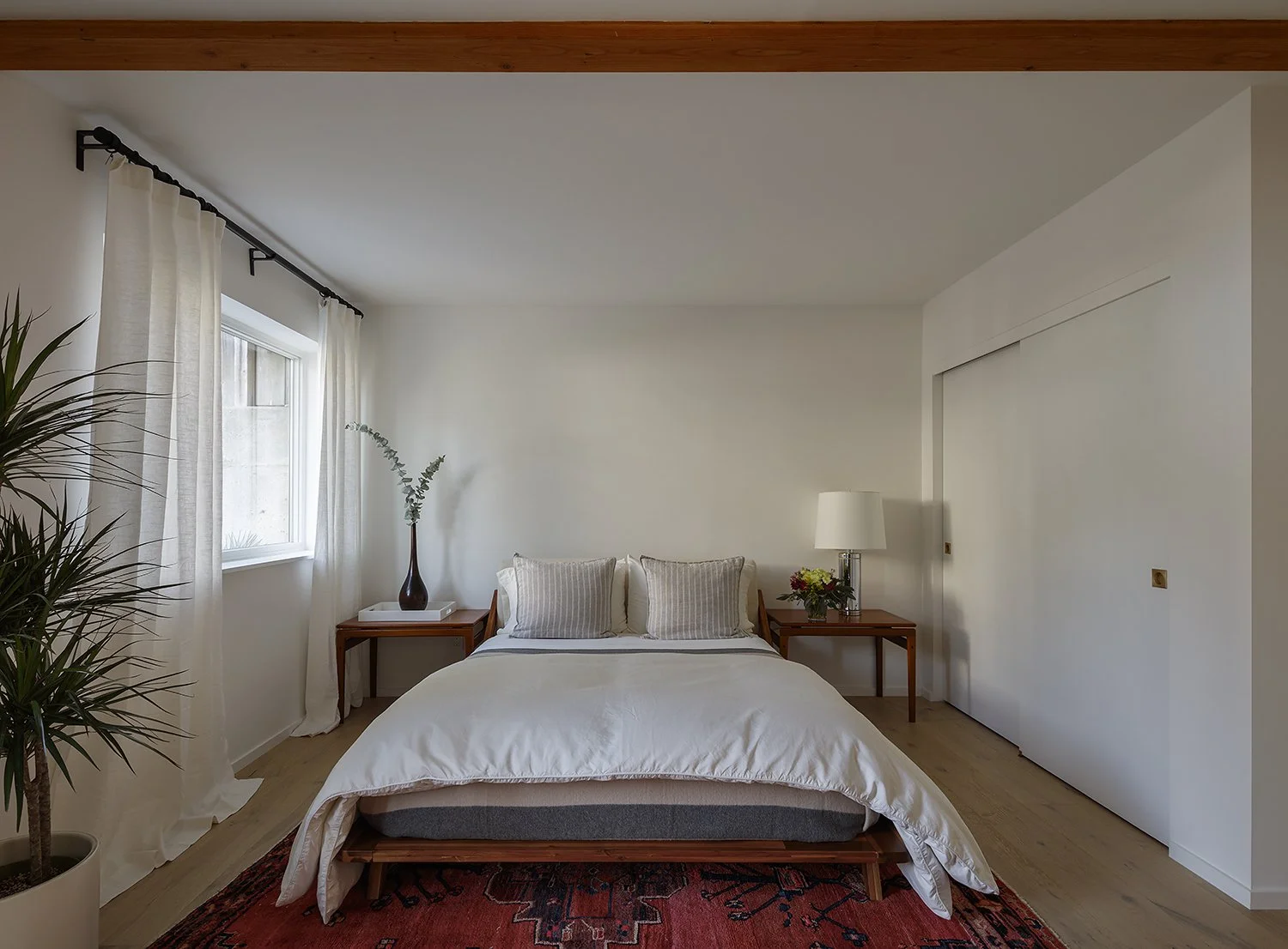 A neatly made bed with two pillows in a bright bedroom. Two wooden nightstands with lamps and flowers, white curtains on a window, and a potted plant in the corner. Hardwood floor with a red patterned rug.