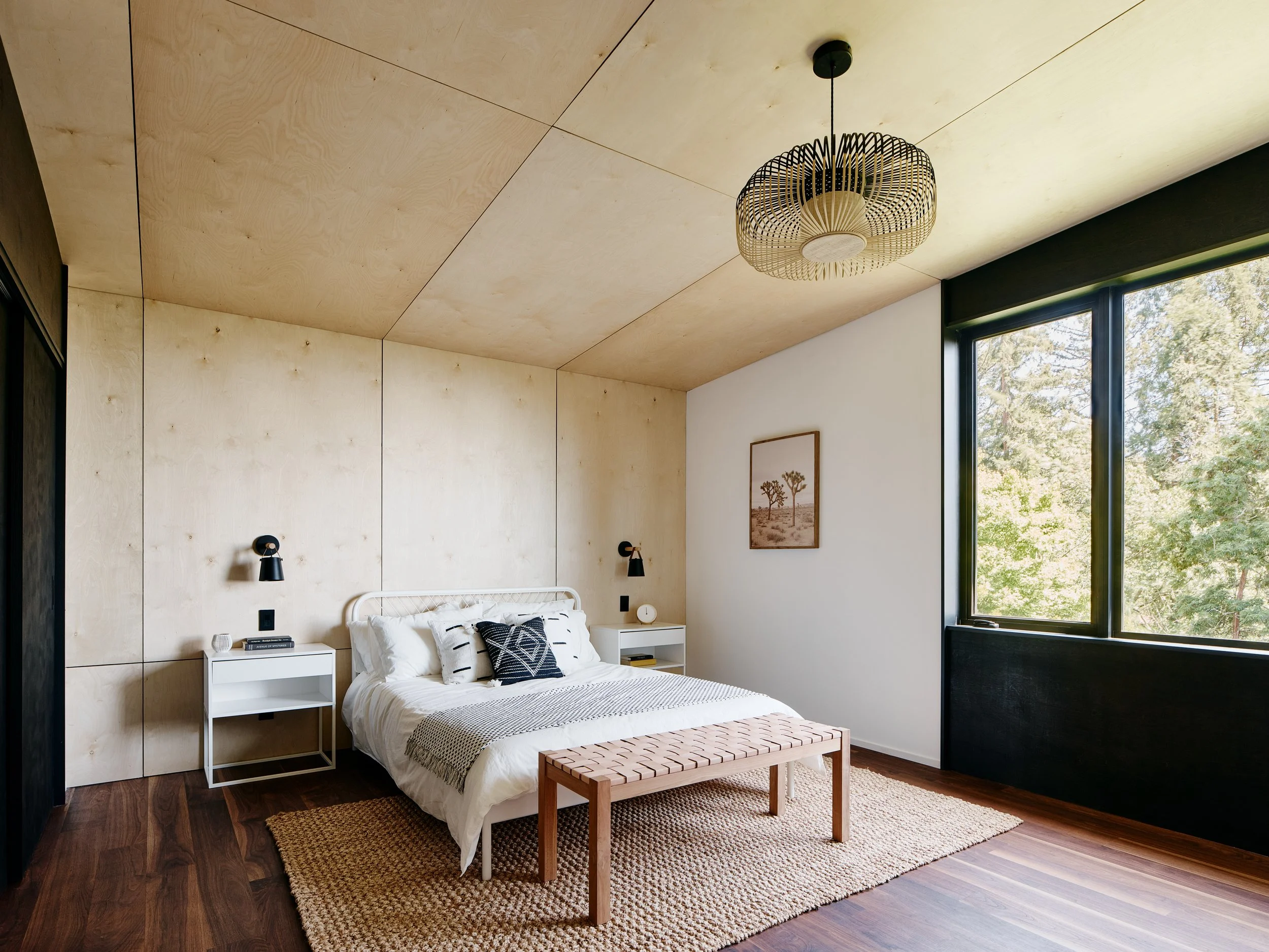 Minimalist bedroom with a white bed, black wall-mounted lights, two white nightstands, a woven bench at the foot of the bed, a framed artwork on the wall, large window with greenery outside, wooden floors, and a textured rug.