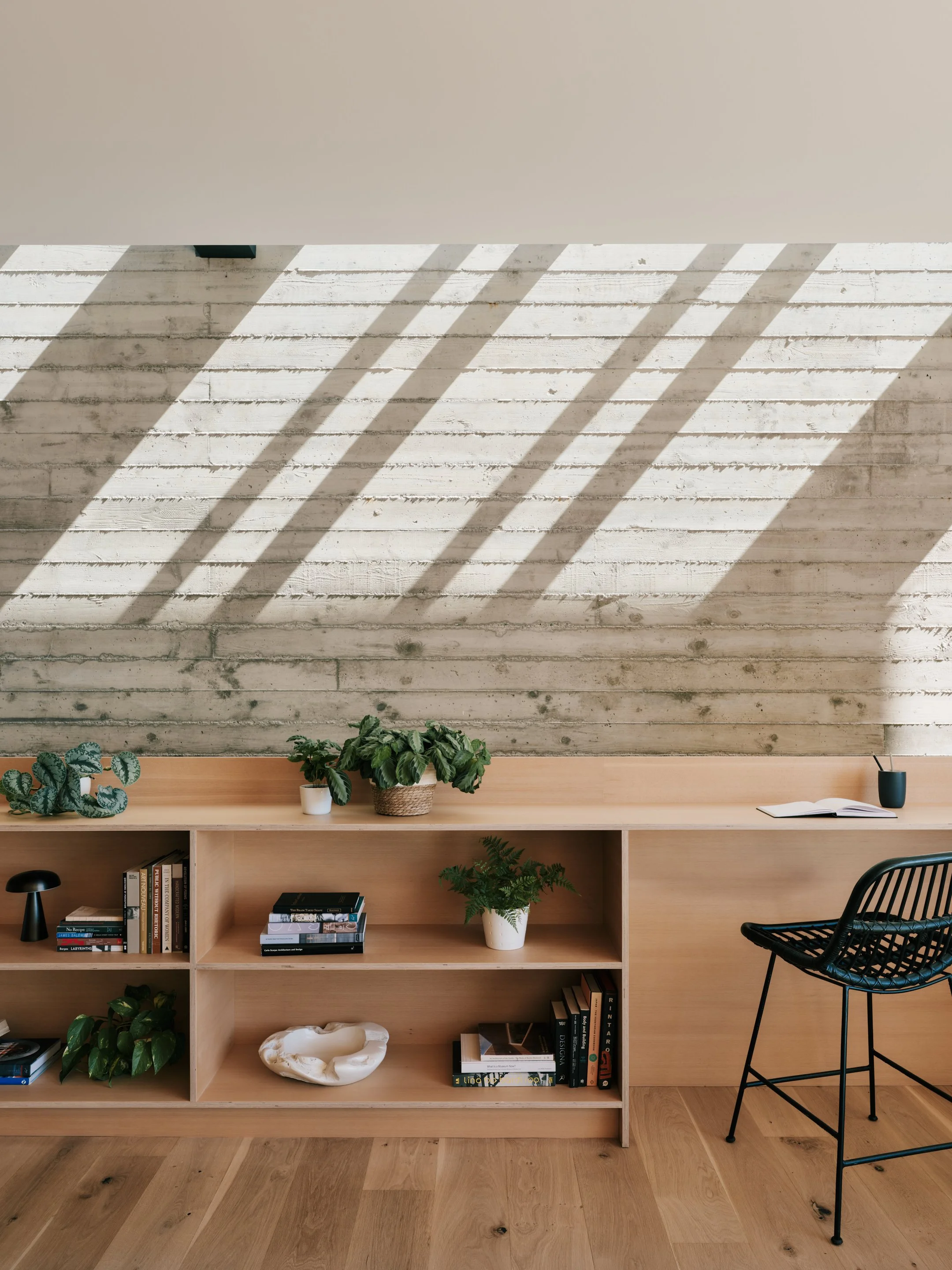 A minimalist wooden bookshelf with potted plants, books, decorative objects, and an open magazine, set against a textured white brick wall with sunlight creating diagonal shadows.