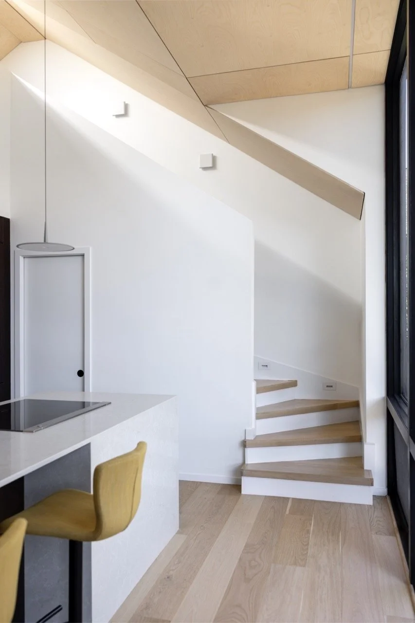 Image of a modern indoor space with wooden flooring, a curved staircase with wooden steps, part of a kitchen with a white countertop, a yellow chair, and large windows.