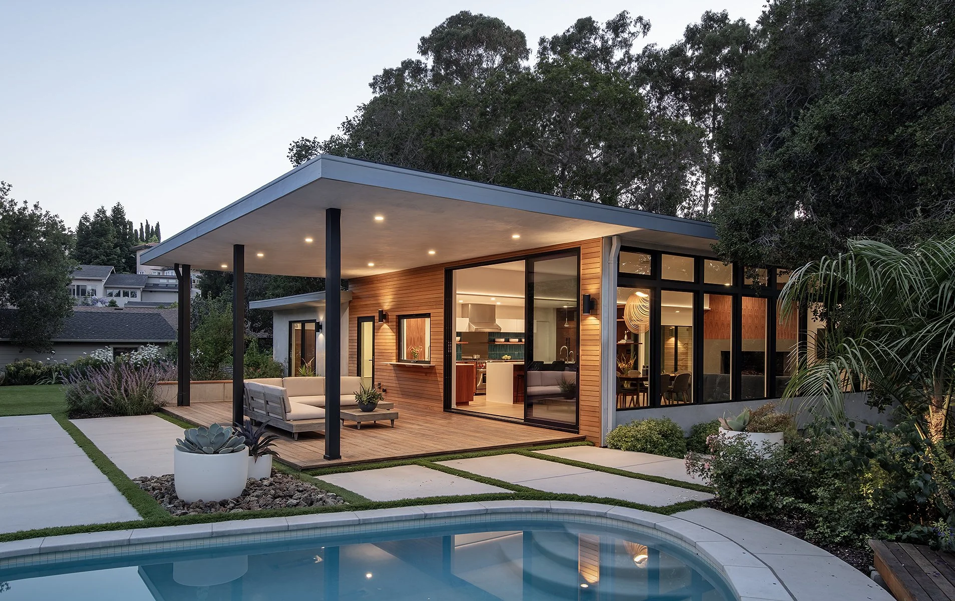 Modern house with large glass windows at dusk, outdoor seating area, swimming pool in the foreground, surrounded by lush greenery.