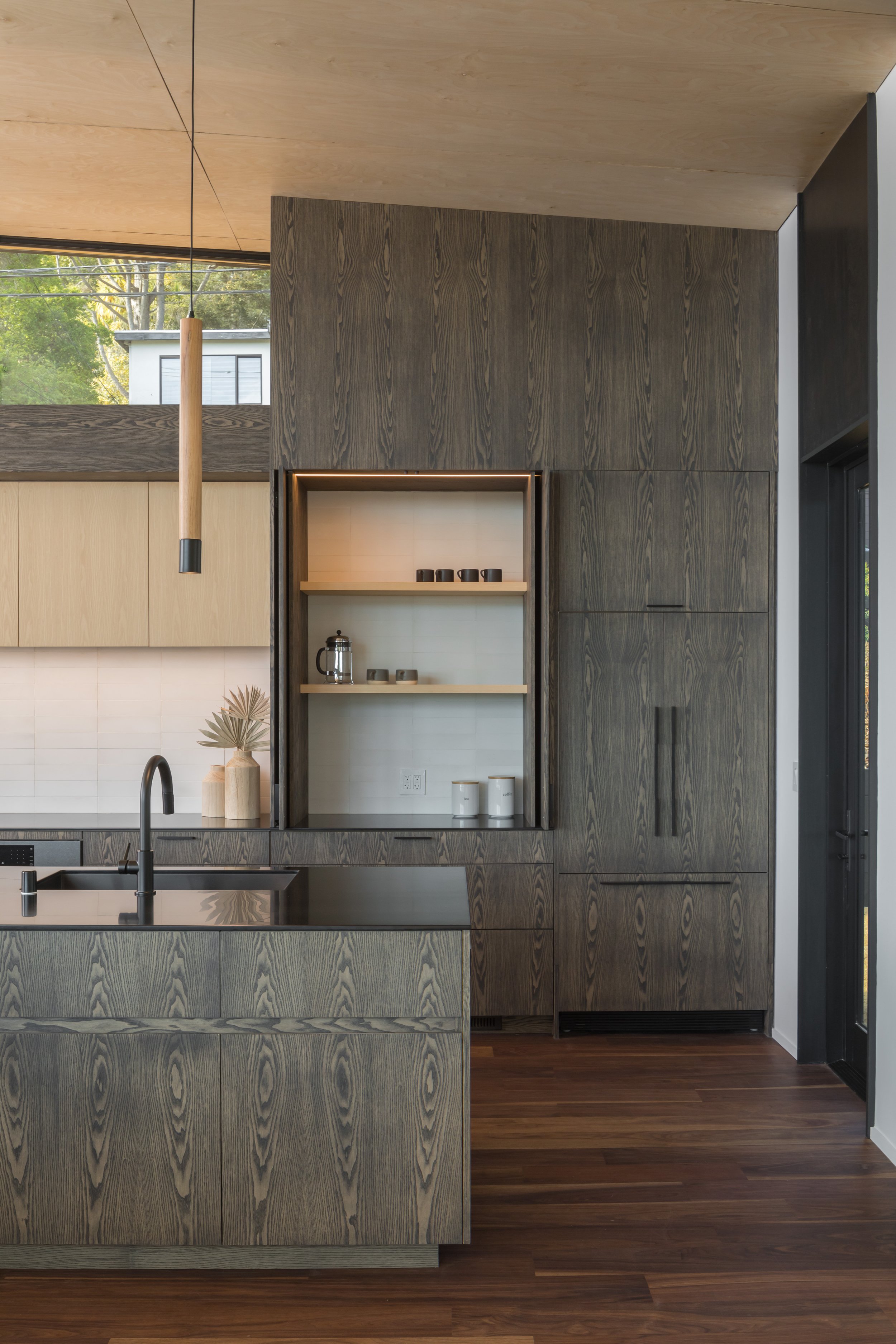 Modern kitchen with dark wood cabinetry, open shelving with dishes, kitchen sink with black faucet, and a hanging wooden pendant light. Large window shows greenery outside.