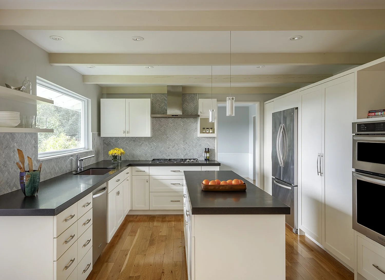 Modern kitchen with white cabinets, black countertops, stainless steel appliances, and light wood flooring. There is a window above the sink and decor including a vase with yellow flowers and a tray of peaches.