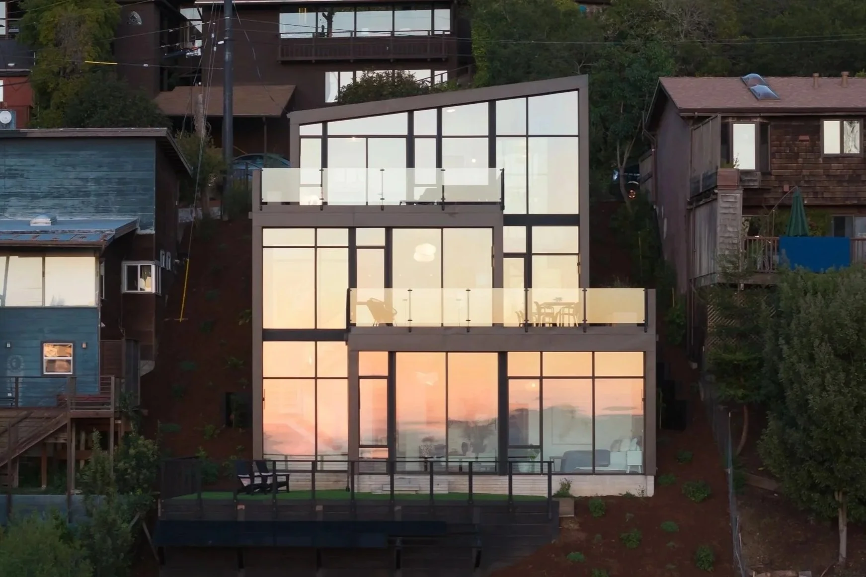 Modern three-story house with large glass windows, illuminated interior, and outdoor balcony, situated on a hillside with neighboring houses surrounding it.