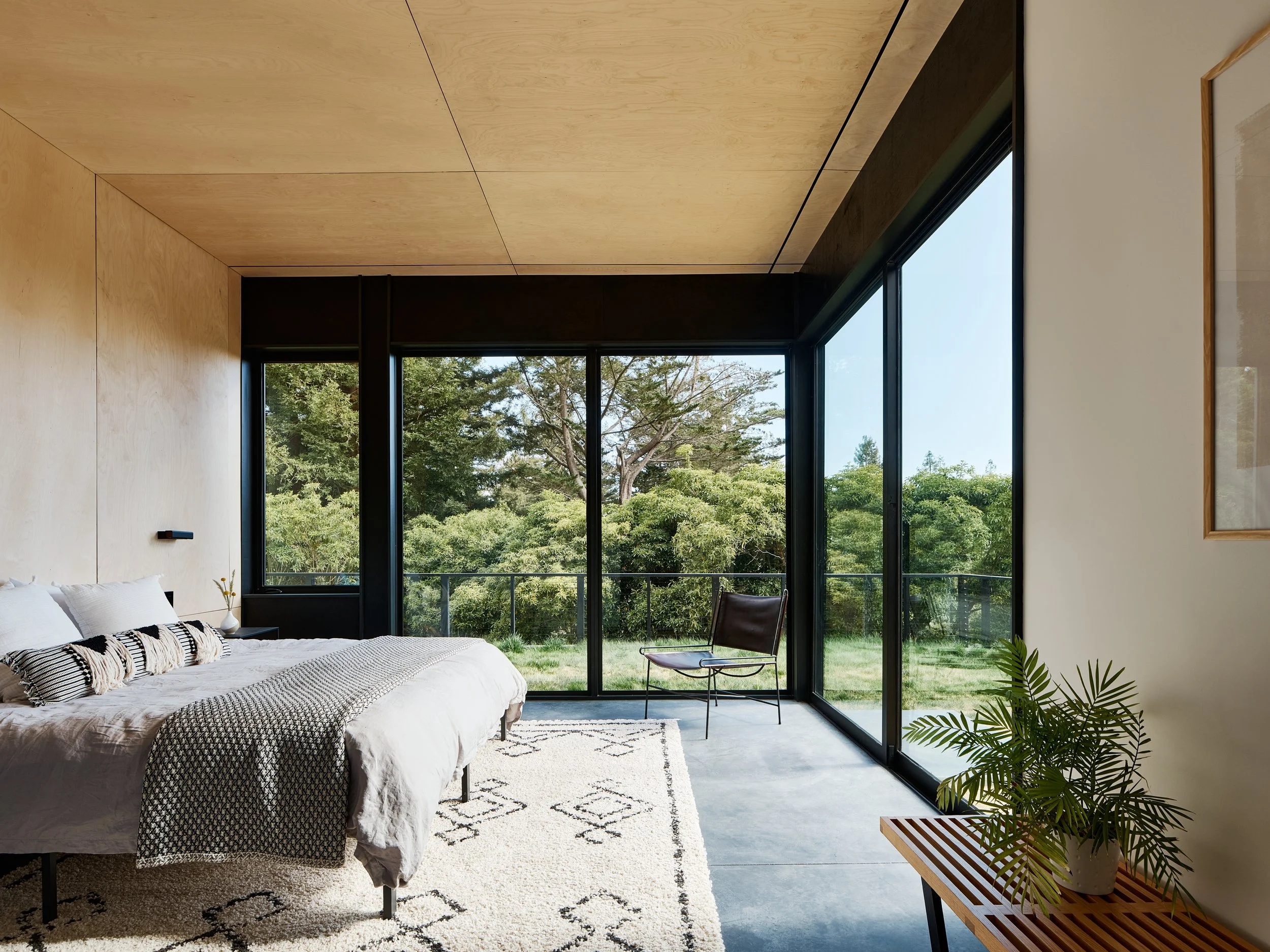 Modern bedroom with large floor-to-ceiling windows showing green trees outside, white bedding with black and white pillows, a beige rug, a wooden bench with plants, and a minimalist aesthetic.