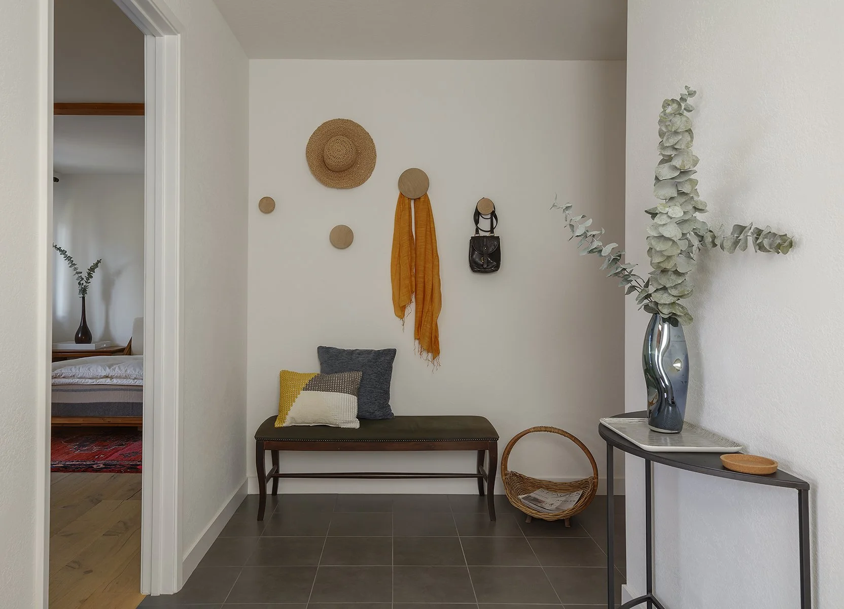 A cozy entryway with a brown bench, gray and yellow pillows, a white wall with decorative hooks holding a hat, scarf, and bag, and a tall vase with eucalyptus on a console table.