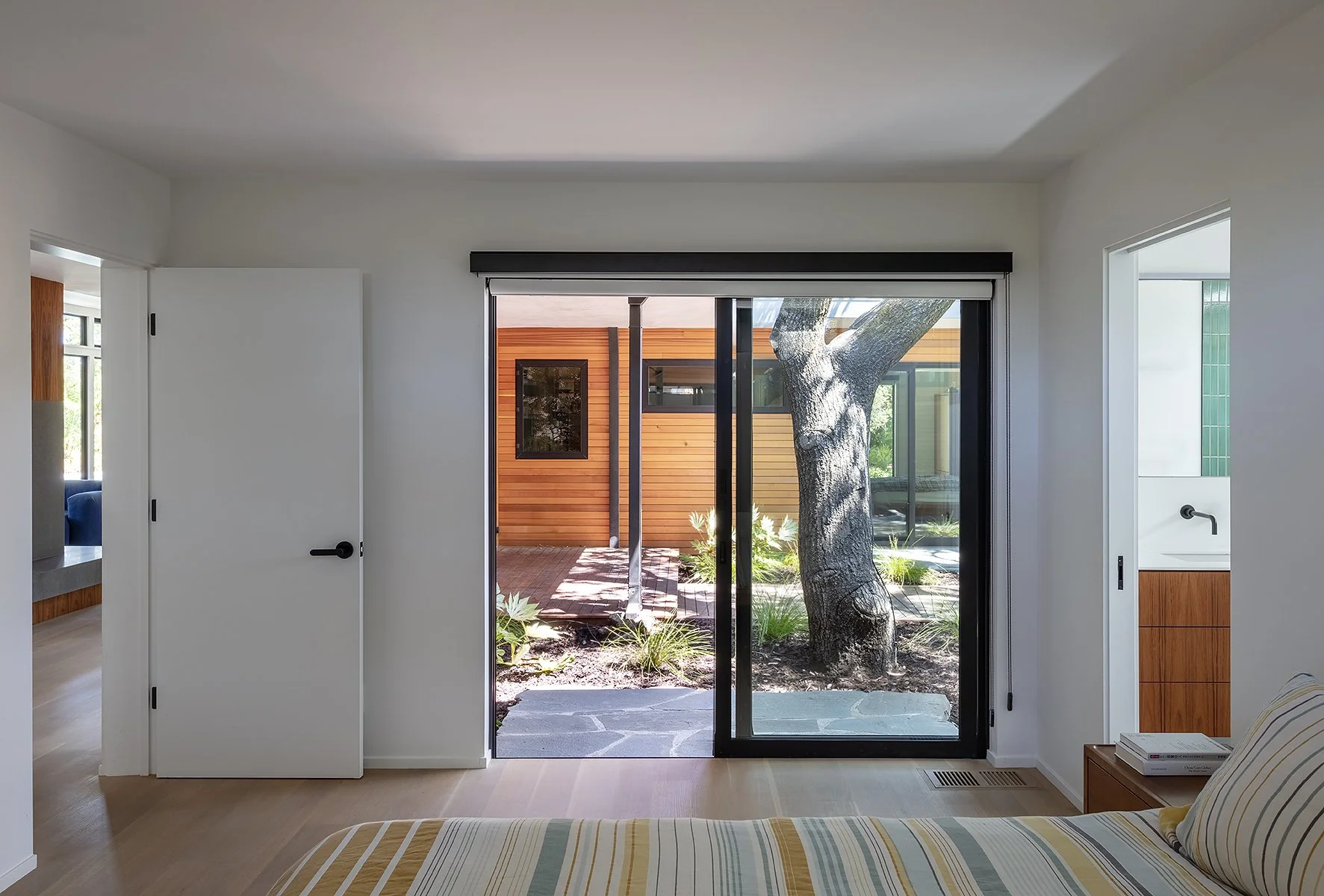 Interior view of a bedroom with sliding glass doors opening to a backyard with a large tree and modern wooden fencing.
