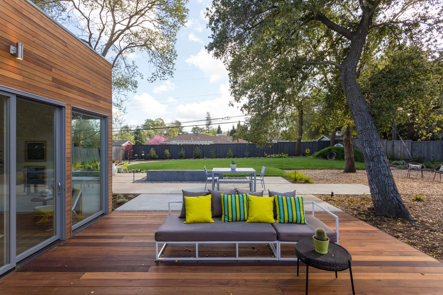 A backyard patio with a wooden deck, a gray sofa with colorful striped and yellow pillows, a small black side table with a potted cactus, and a view of a grassy yard with trees, a fence, and outdoor furniture.