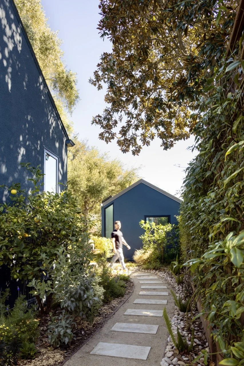 A person walking along a stone pathway in a lush garden with blue houses and dense green foliage on a sunny day.