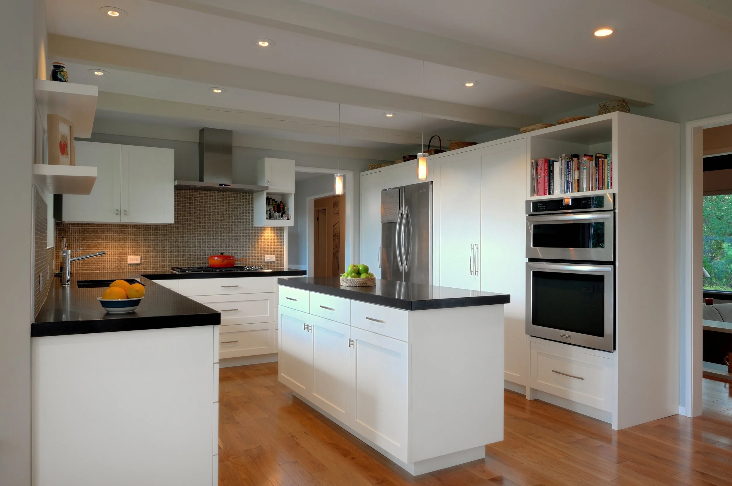 Modern white kitchen with island, stainless steel appliances, and warm wooden floors, decorated with bowls of fruit.