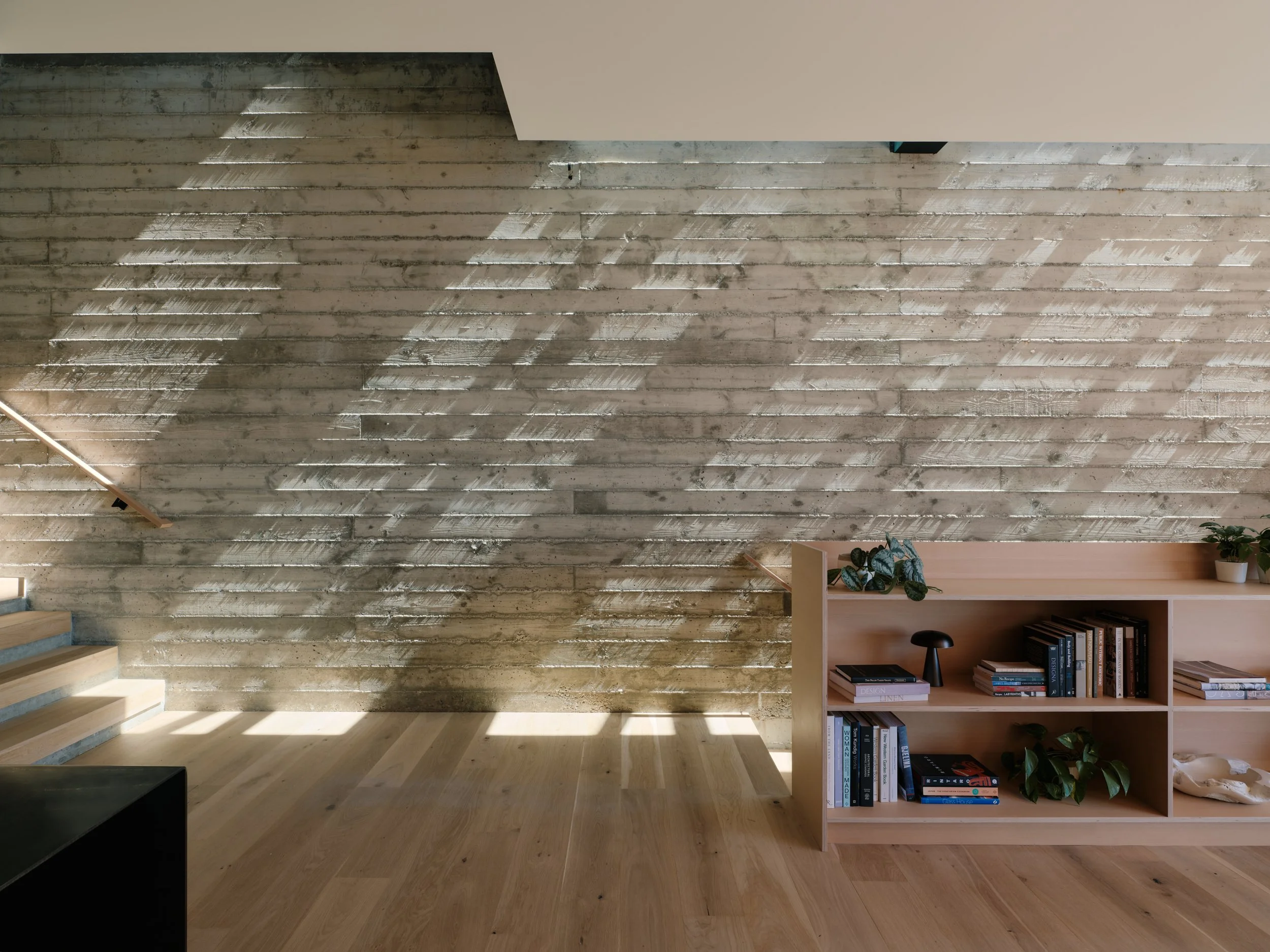 A modern interior with a wooden staircase, a concrete wall with diagonal shadows, and a bookshelf filled with books and plants.