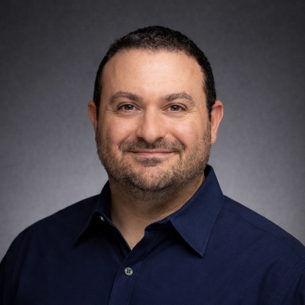 Headshot of a man with dark hair, beard, wearing a dark blue collared shirt, smiling, against a gray background.