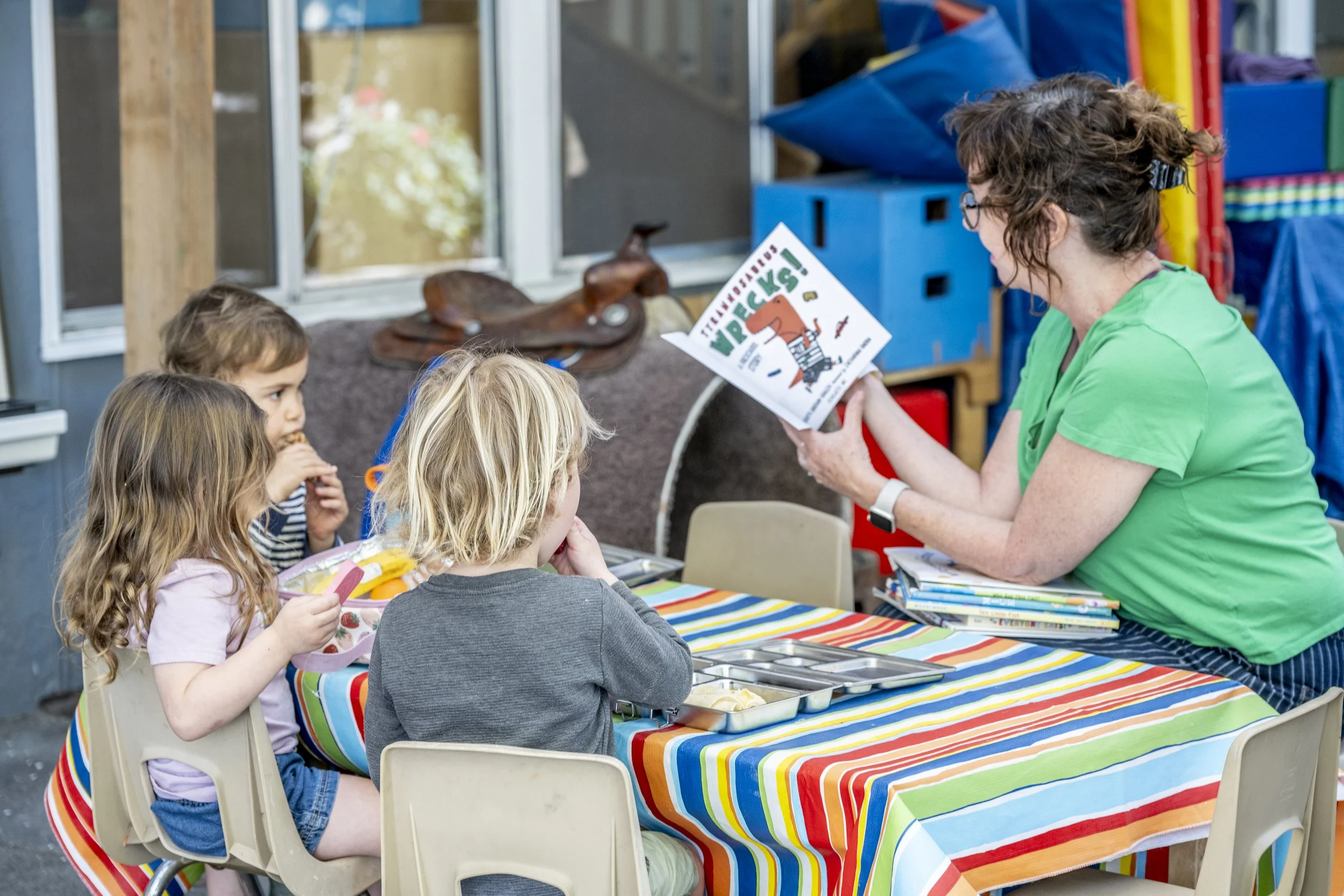 Teacher Chandra outside at a table, reading a book to students while they eat a snack