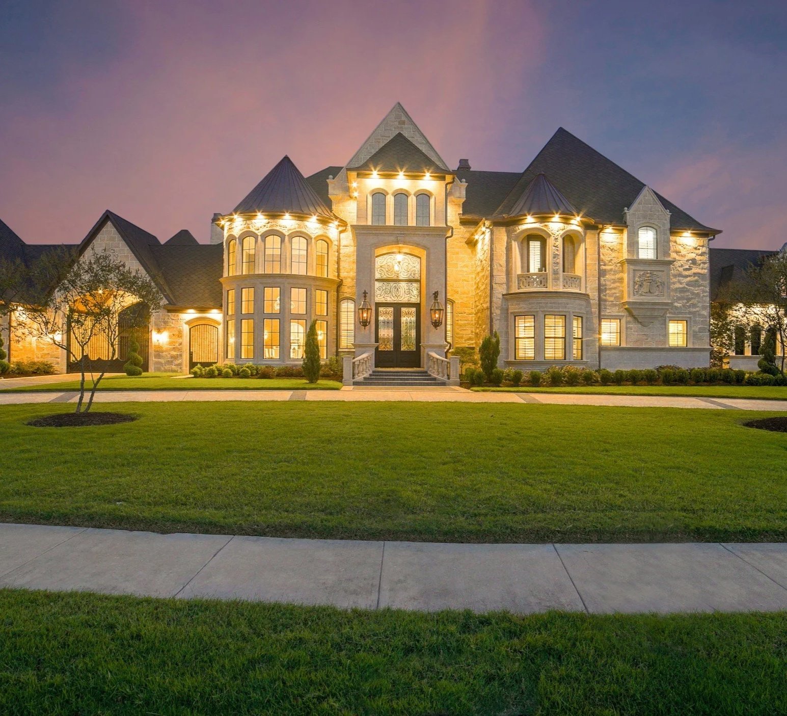 A large, elegant mansion with stone exterior lit up at dusk, surrounded by a well-manicured lawn and a sidewalk.