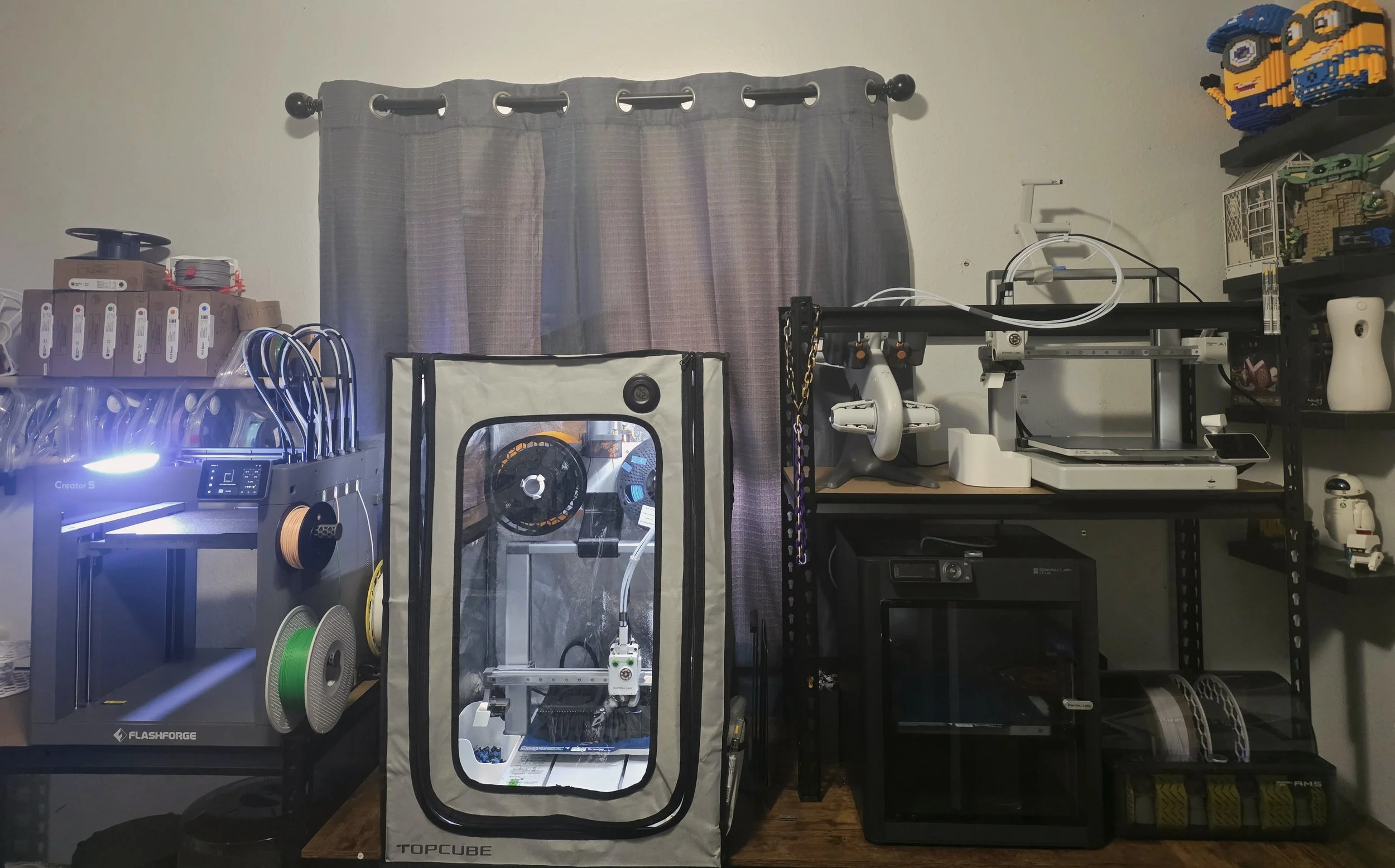 A room with various 3D printing and maker equipment, including a 3D printer, filament spools, a print enclosure, and miniature LEGO creations on shelves.
