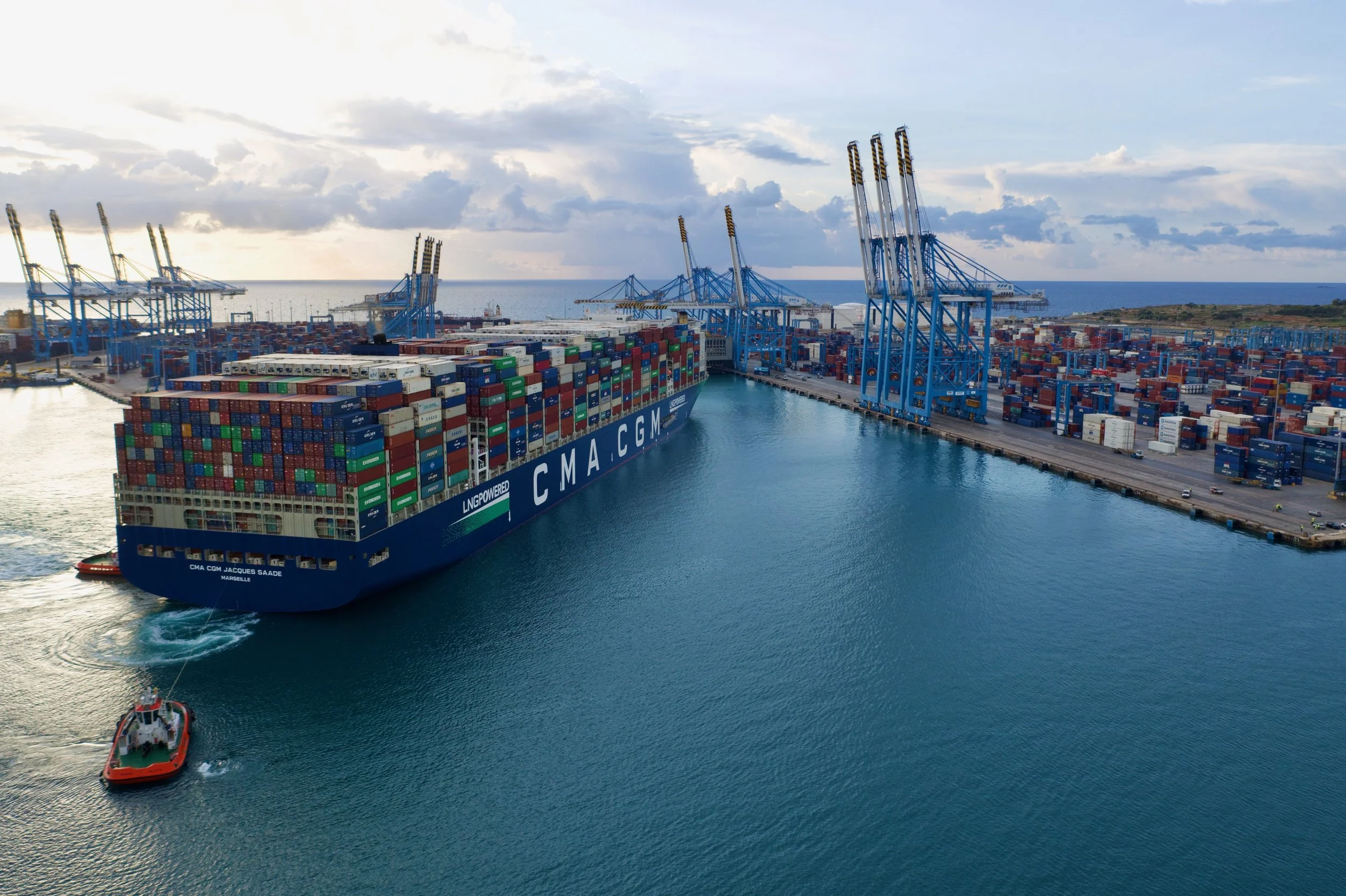 A large cargo ship loaded with stacked shipping containers is leaving a port, accompanied by a small tugboat. The port has blue cranes for loading and unloading containers with numerous containers stacked on land, with an ocean and cloudy sky in the background.