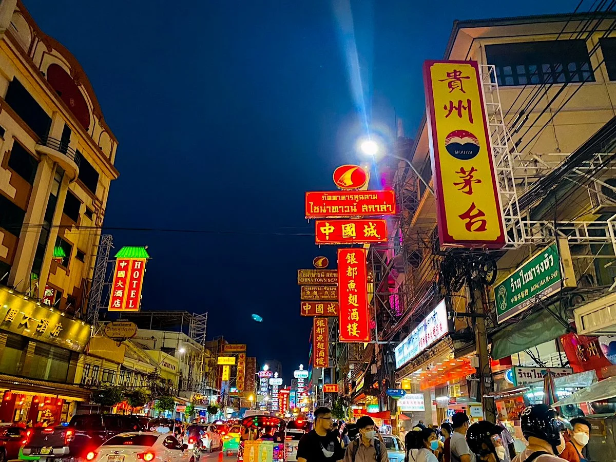 Nighttime street scene with colorful neon signs in Chinese and Thai characters, crowded with pedestrians wearing face masks and vehicles on the busy road.