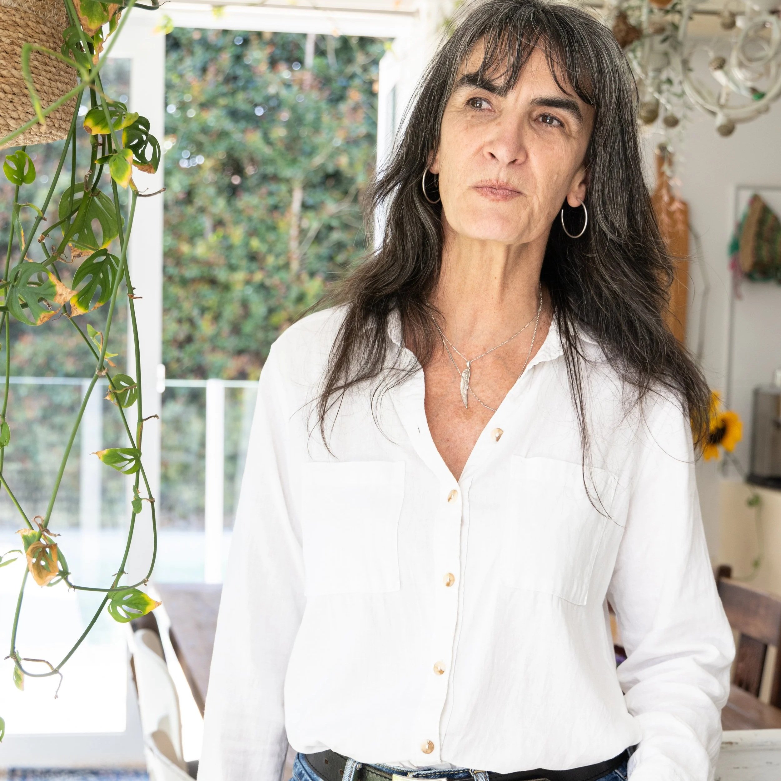 Close-up of a woman with long black hair, wearing hoop earrings and a white shirt, standing indoors near a window with a leafy outdoor scene in the background.