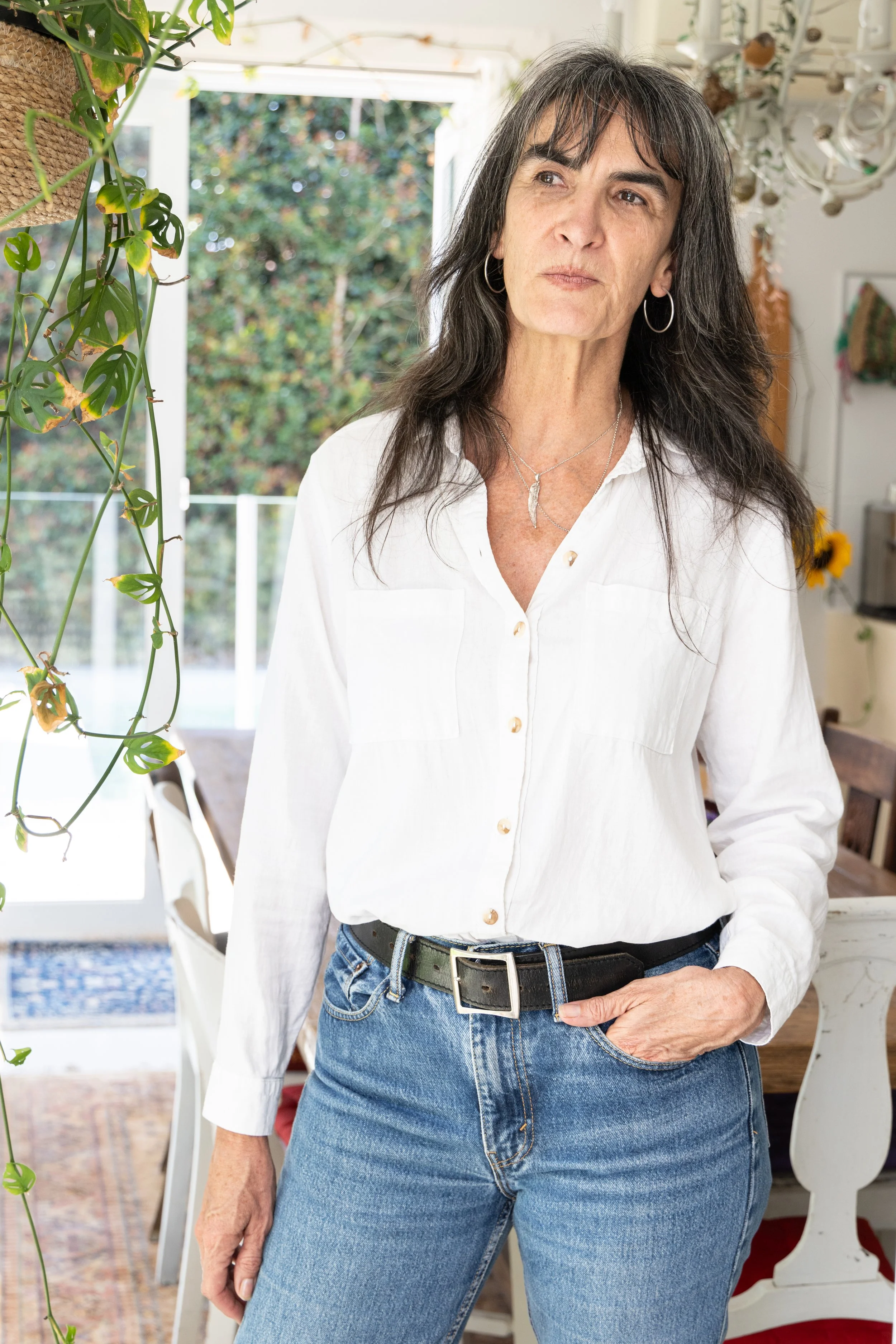 A woman with long dark hair, wearing a white button-up shirt and blue jeans, stands indoors near a window with plants and decor in the background.