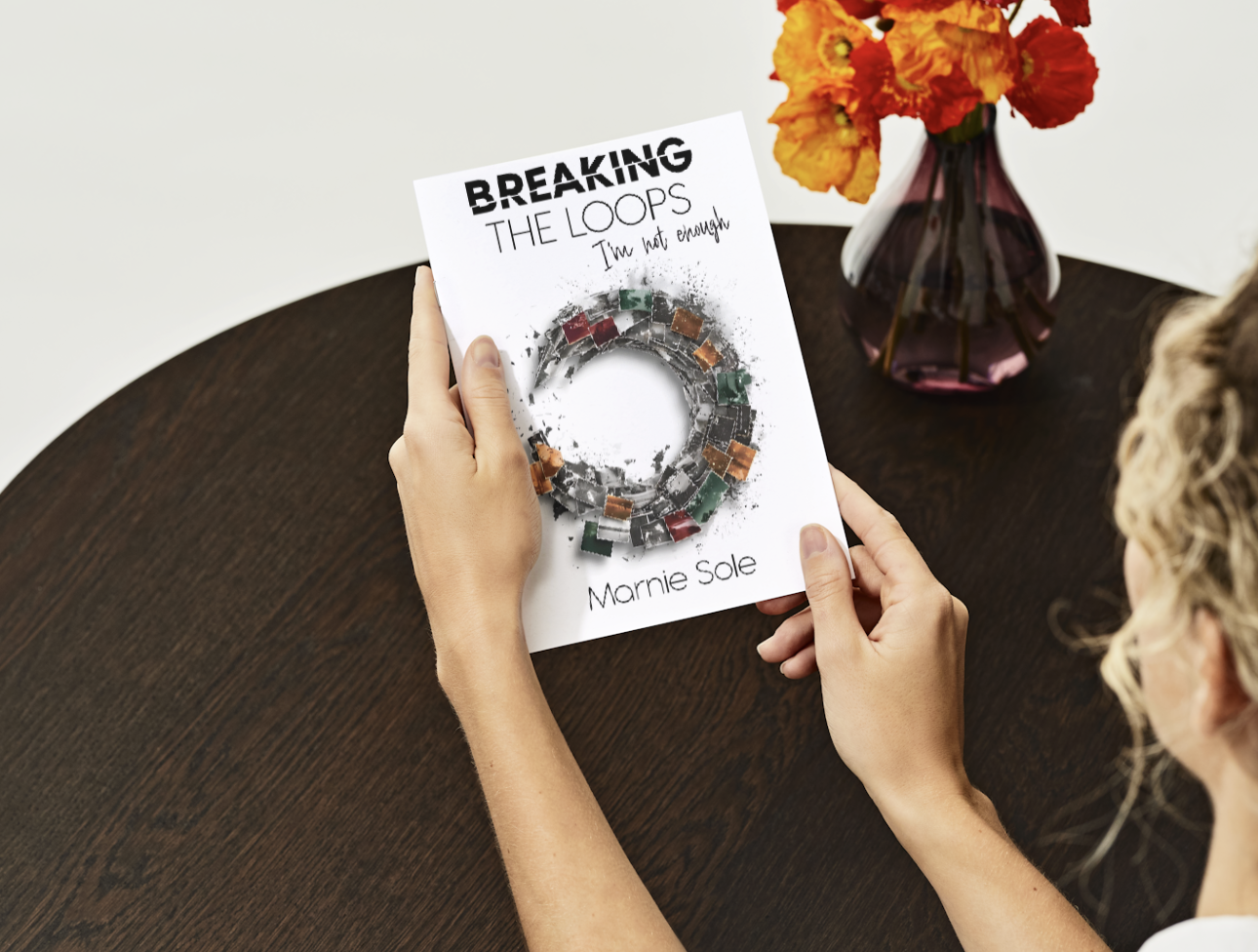 A person holding a book titled 'Breaking the Loops: I'm not enough' by Marnie Sole. The book features an illustration of a circular mechanical loop on the cover. The person is sitting at a dark wooden table with a vase of orange and yellow flowers in the background.