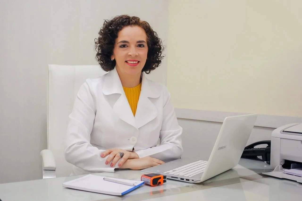 Mulher sorridente com cabelo cacheado, usando jaleco branco, sentada em mesa de escritório com laptop, caderno, caneta, e impressora ao fundo.