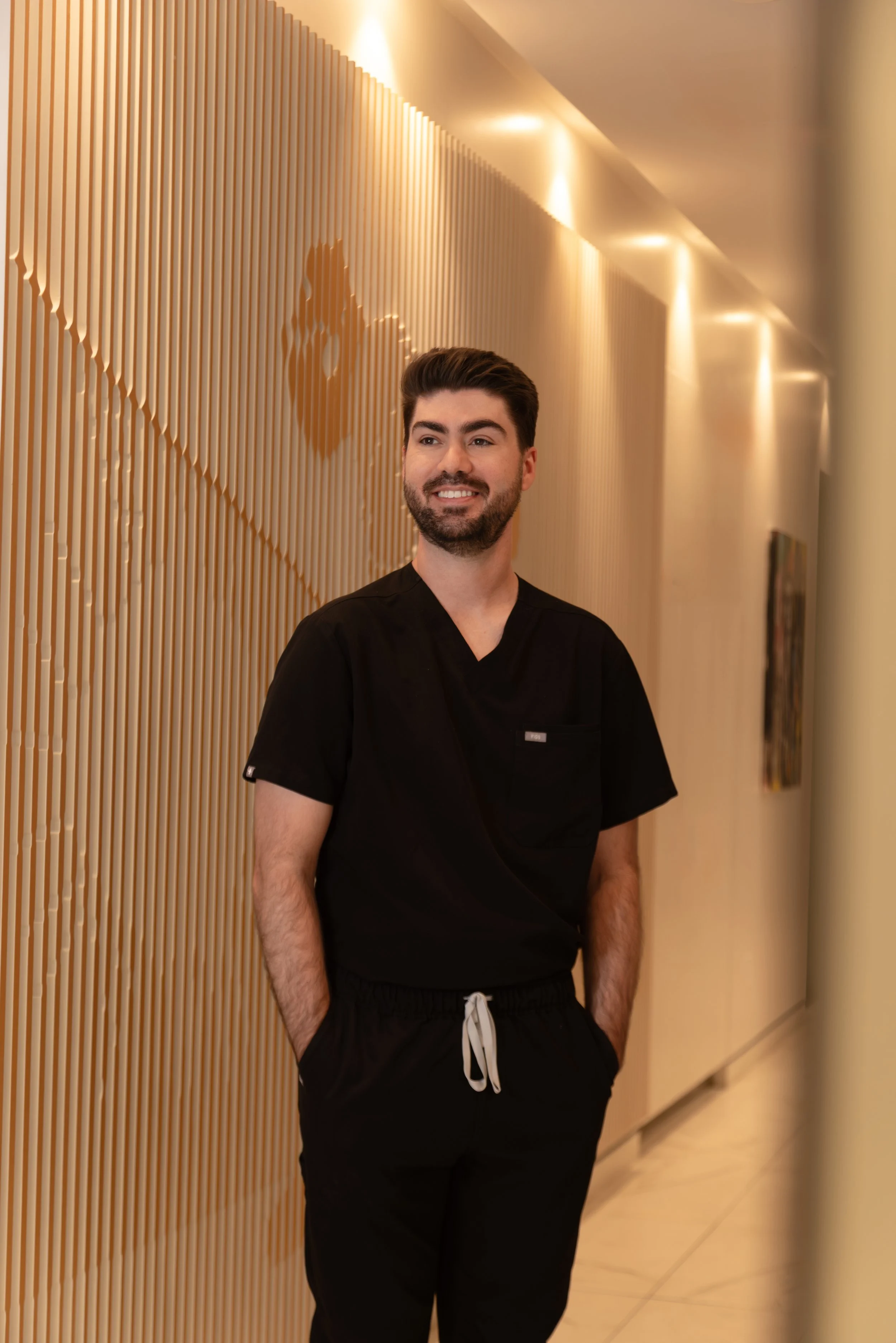 A smiling man wearing black scrubs standing in a hallway with wooden paneling on the wall.