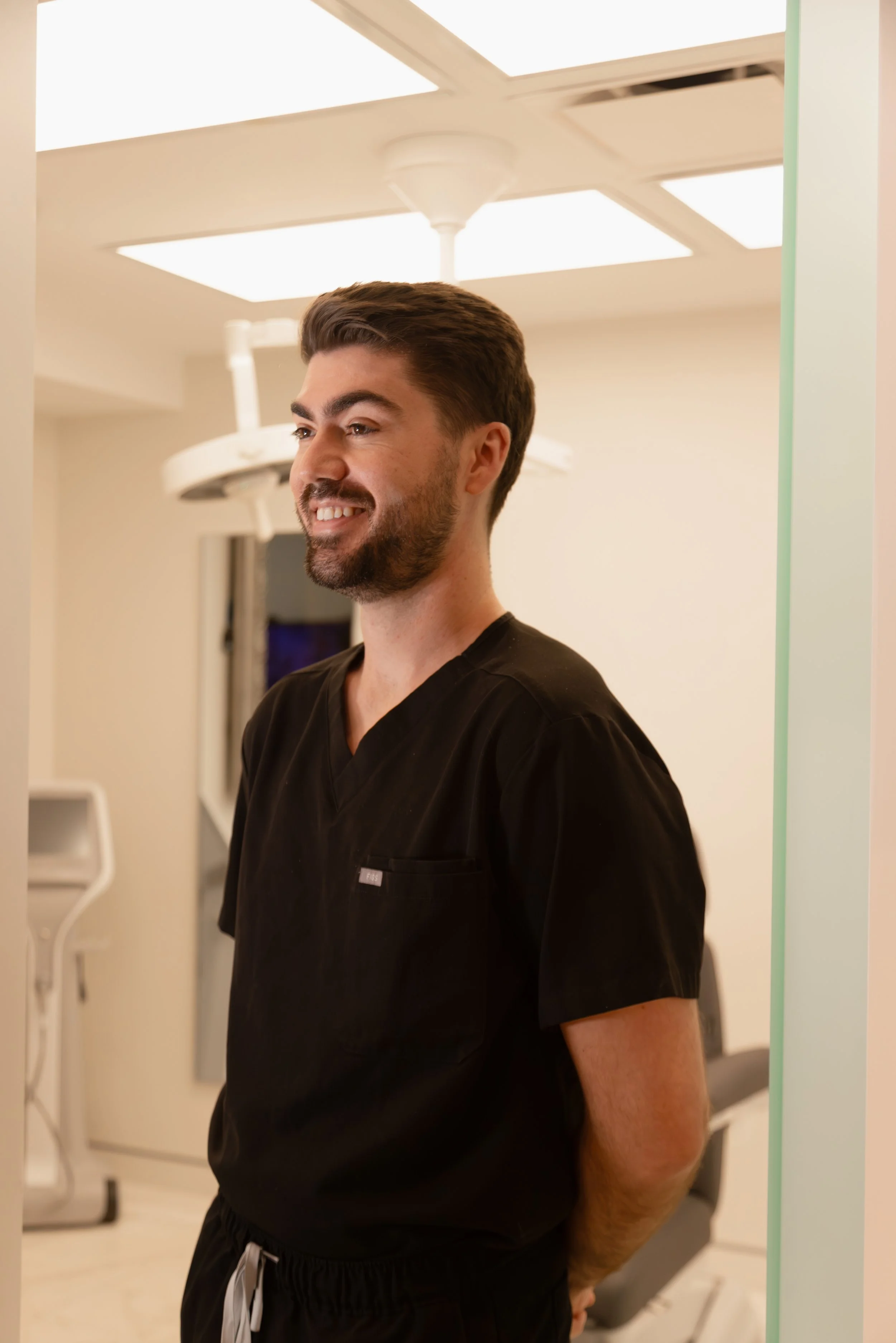 A male healthcare professional in black scrubs standing in a medical facility with a bright, clinical environment and medical equipment in the background.