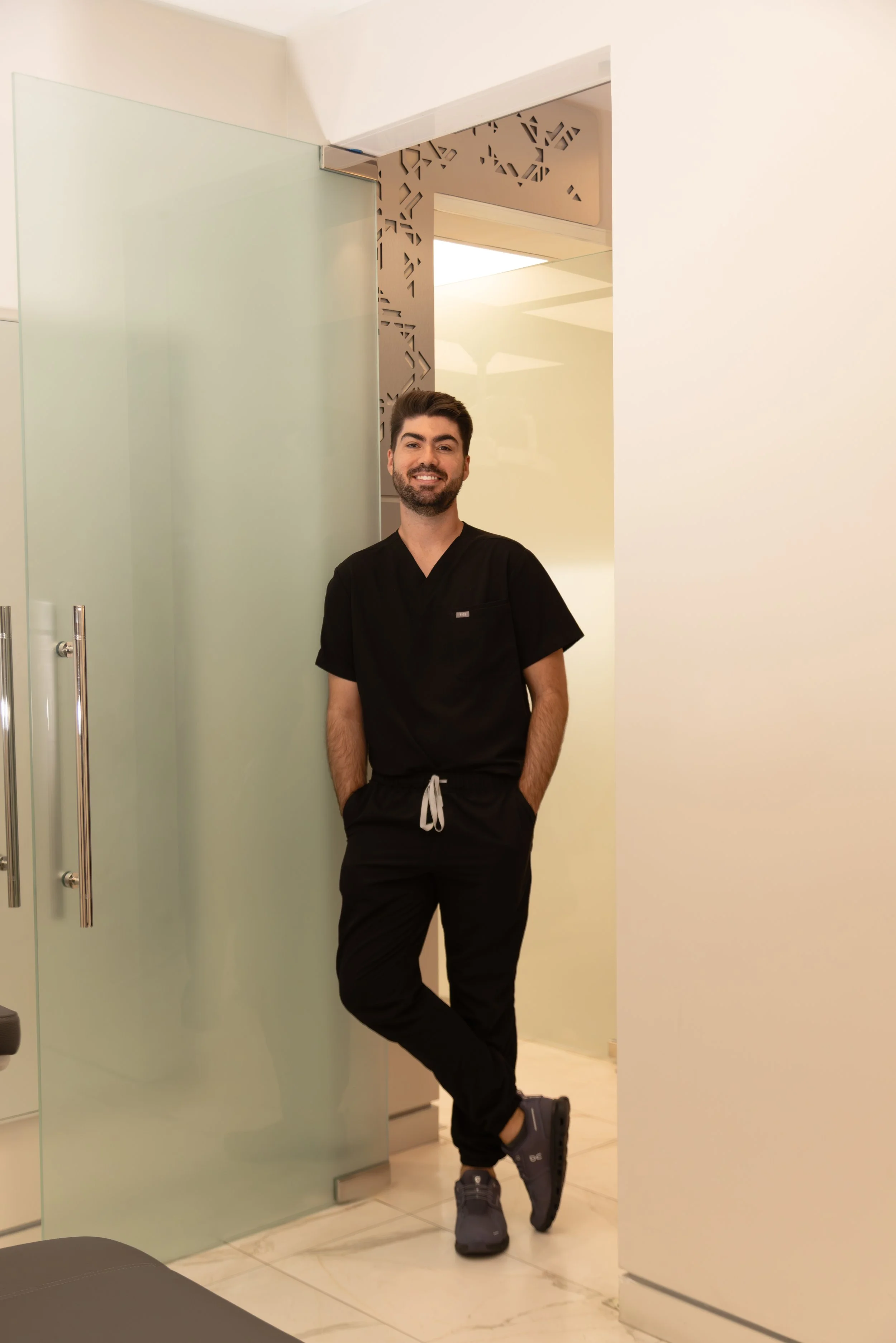 A man with dark hair, a beard, and a big smile wearing black scrubs and sneakers, standing and leaning against a glass door in a modern, minimalist room.