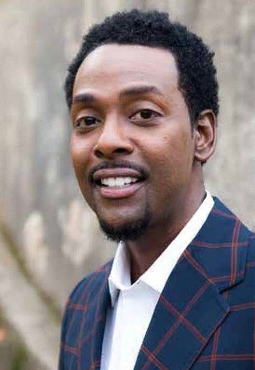 A portrait of a man with short curly hair, a mustache, and a goatee, wearing a white shirt and a dark blue plaid blazer in an outdoor setting with a wooden background.
