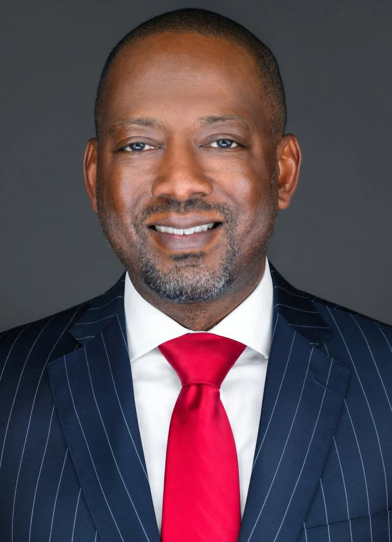 Professional headshot of a smiling African American man in a navy pinstripe suit, white shirt, and red tie against a dark grey background.
