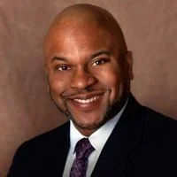 Portrait of a smiling Black man in a suit and tie against a brown background.