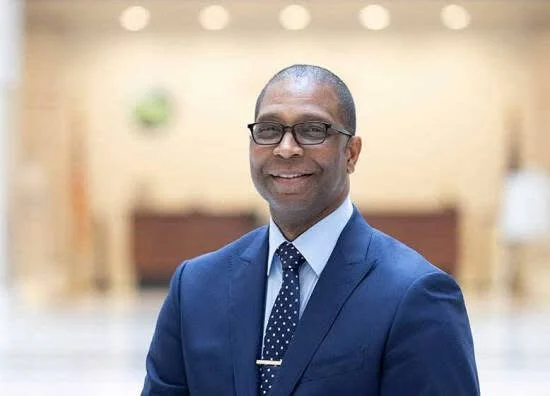 Man in a blue suit, glasses, and tie smiling in an indoor setting.