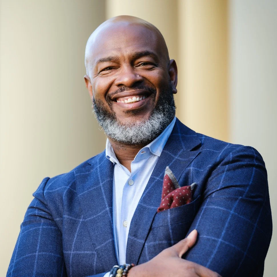 A smiling man with a beard and shaved head, wearing a blue checkered blazer, a light blue shirt, and a red pocket square, standing outdoors in front of a neutral background.
