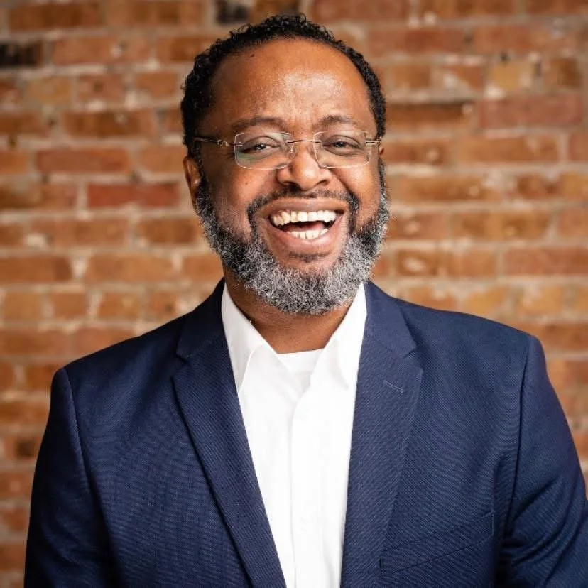 Smiling man in glasses and a navy suit, with a beard and gray hair, standing in front of a brick wall.