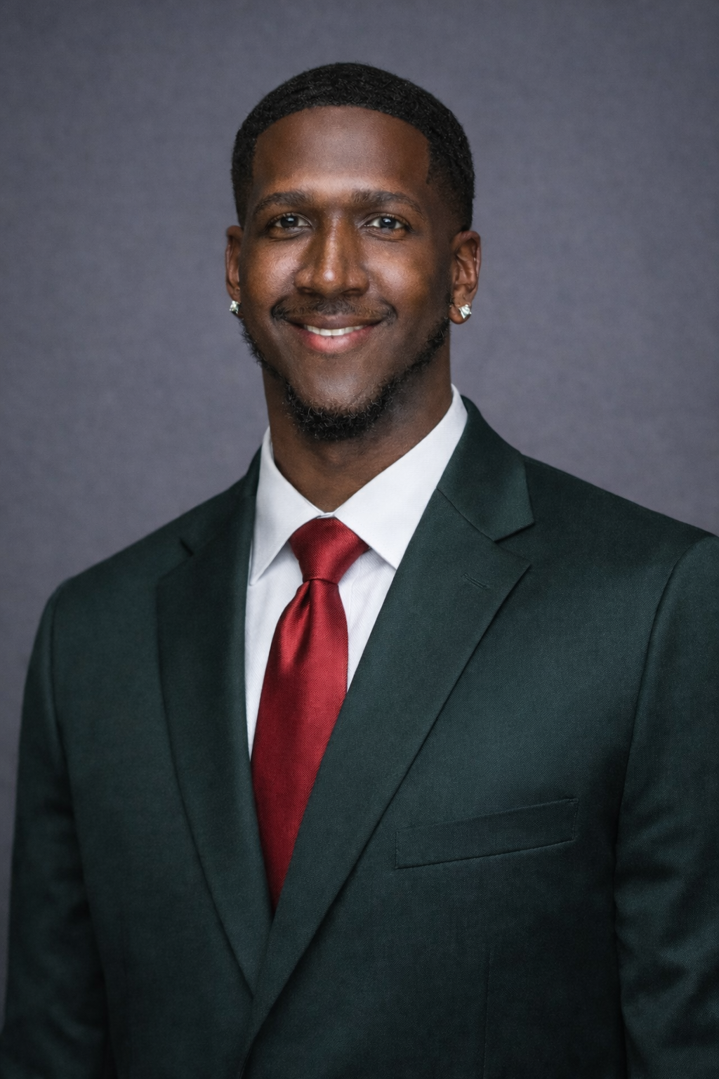 A young man in a dark suit, white shirt, and red tie, smiling, with earrings, against a plain dark background.