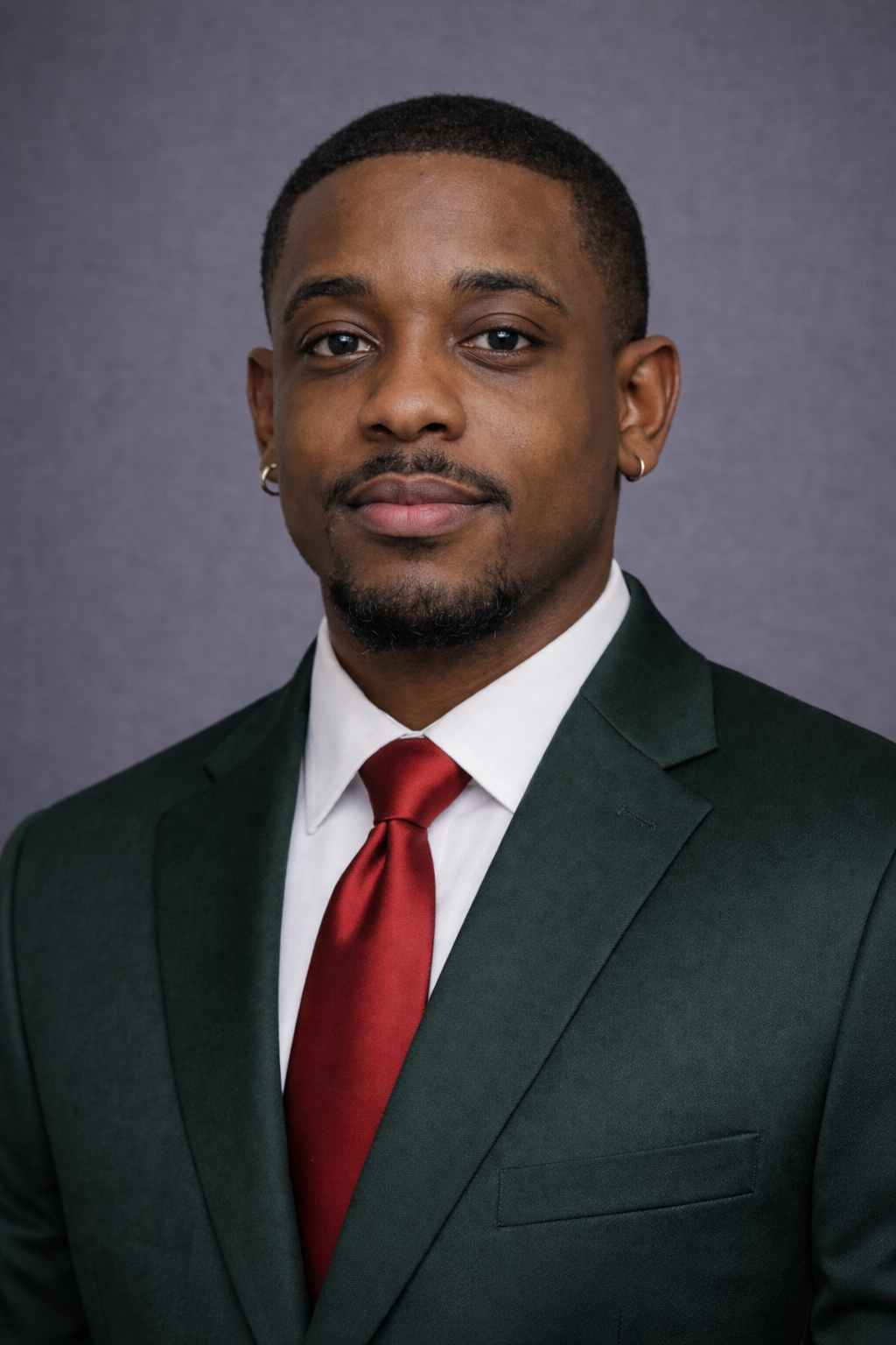 A young Black man in a dark green suit with a white shirt and red tie, posing against a gray background.