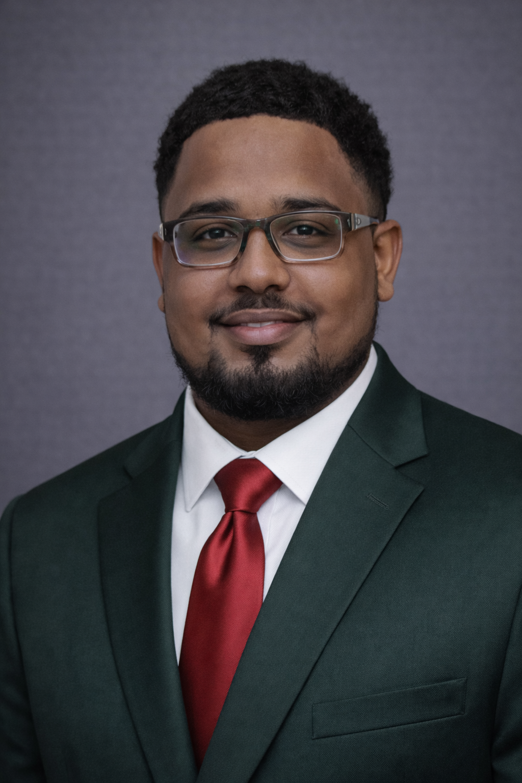 Formal portrait of a young man with glasses, wearing a dark suit, white shirt, and red tie, against a plain background.