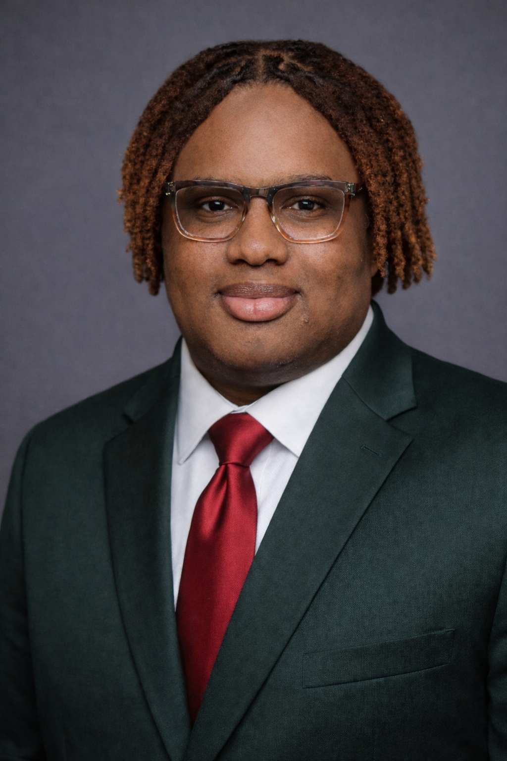 Portrait of a young man with glasses, wearing a dark suit, white shirt, and red tie, against a gray background.