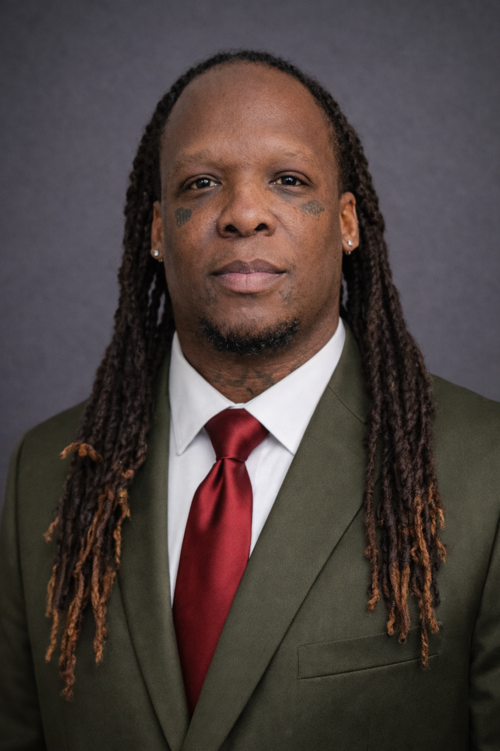 Professional portrait of a man with long dreadlocks wearing a suit and red tie against a gray background.
