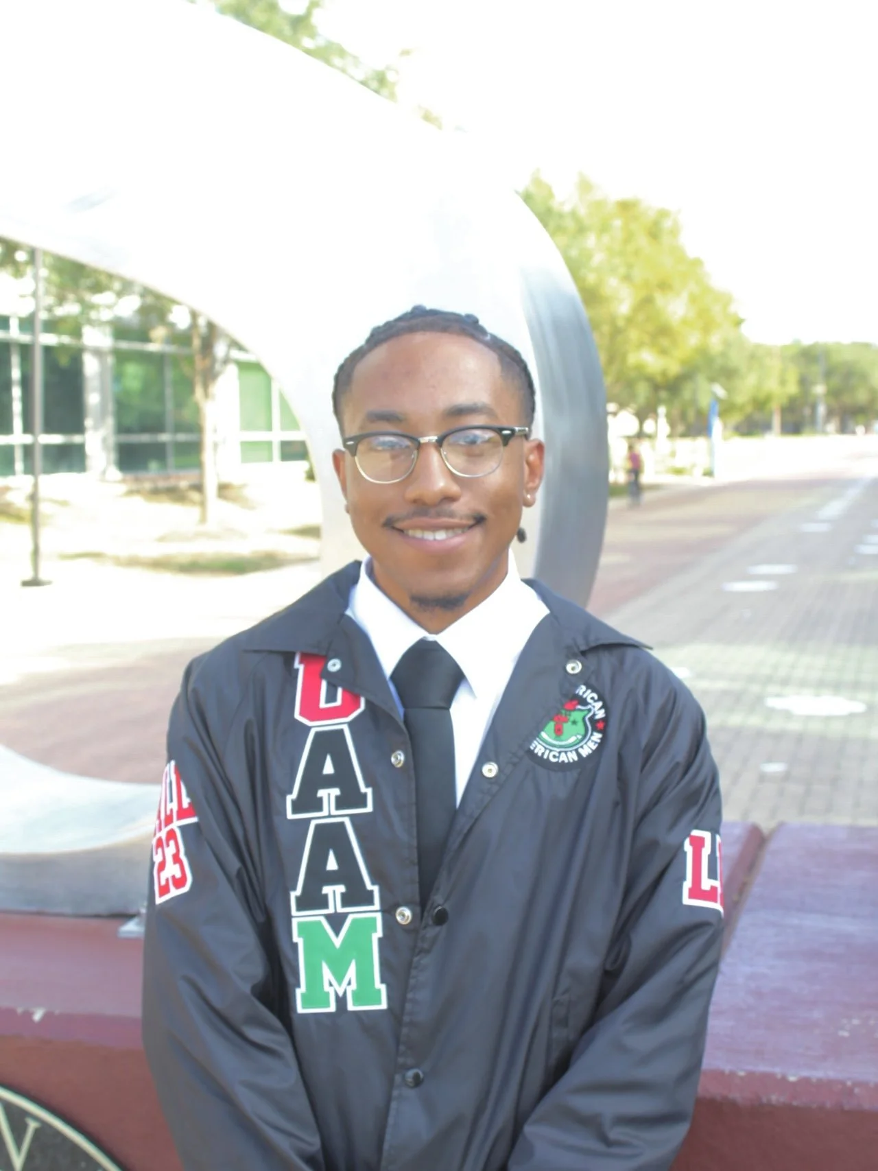 A young man with glasses and a goatee, smiling, wearing a black jacket with colorful letters and embroidery, standing outdoors near a modern sculpture and trees.
